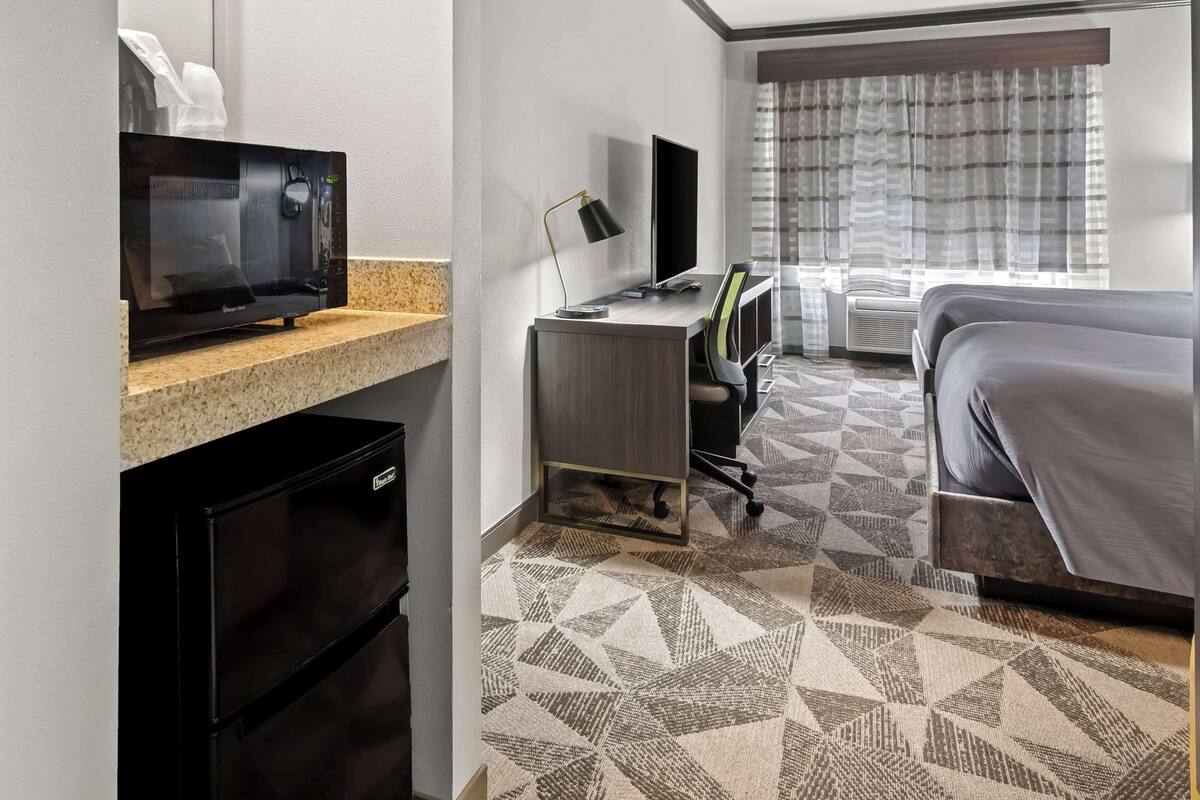 Quarto de hotel com cozinha compacta, uma cama, escrivaninha com computador e uma janela grande com cortinas.