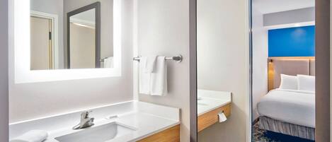 A modern bathroom with a large mirror, two sinks, and a visible door leading to a bedroom with a bed and a blue accent wall.