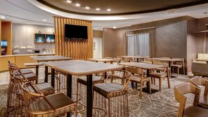 Un comedor de hotel con varias mesas y sillas, un televisor montado en un panel de madera, y una zona de bar con una caja registradora.