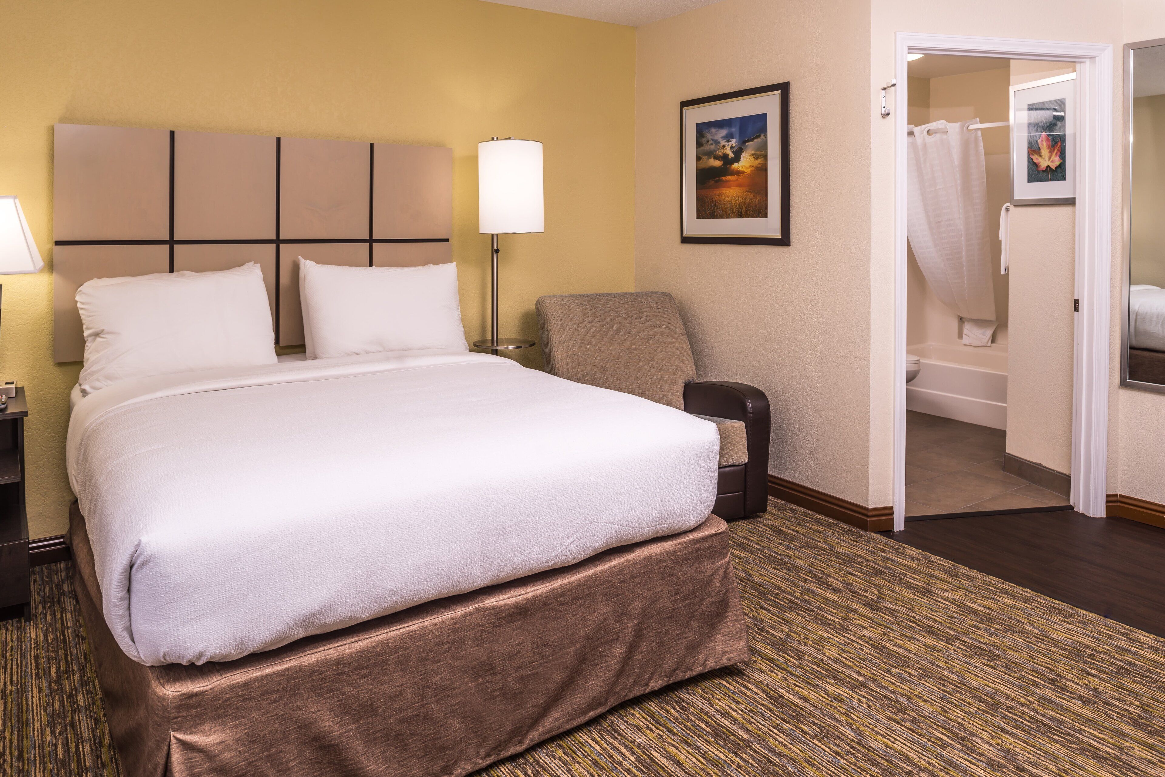 Quarto de hotel com uma cama grande, uma cadeira, uma mesinha de cabeceira, uma luminária, um quadro na parede e a porta do banheiro aberta.