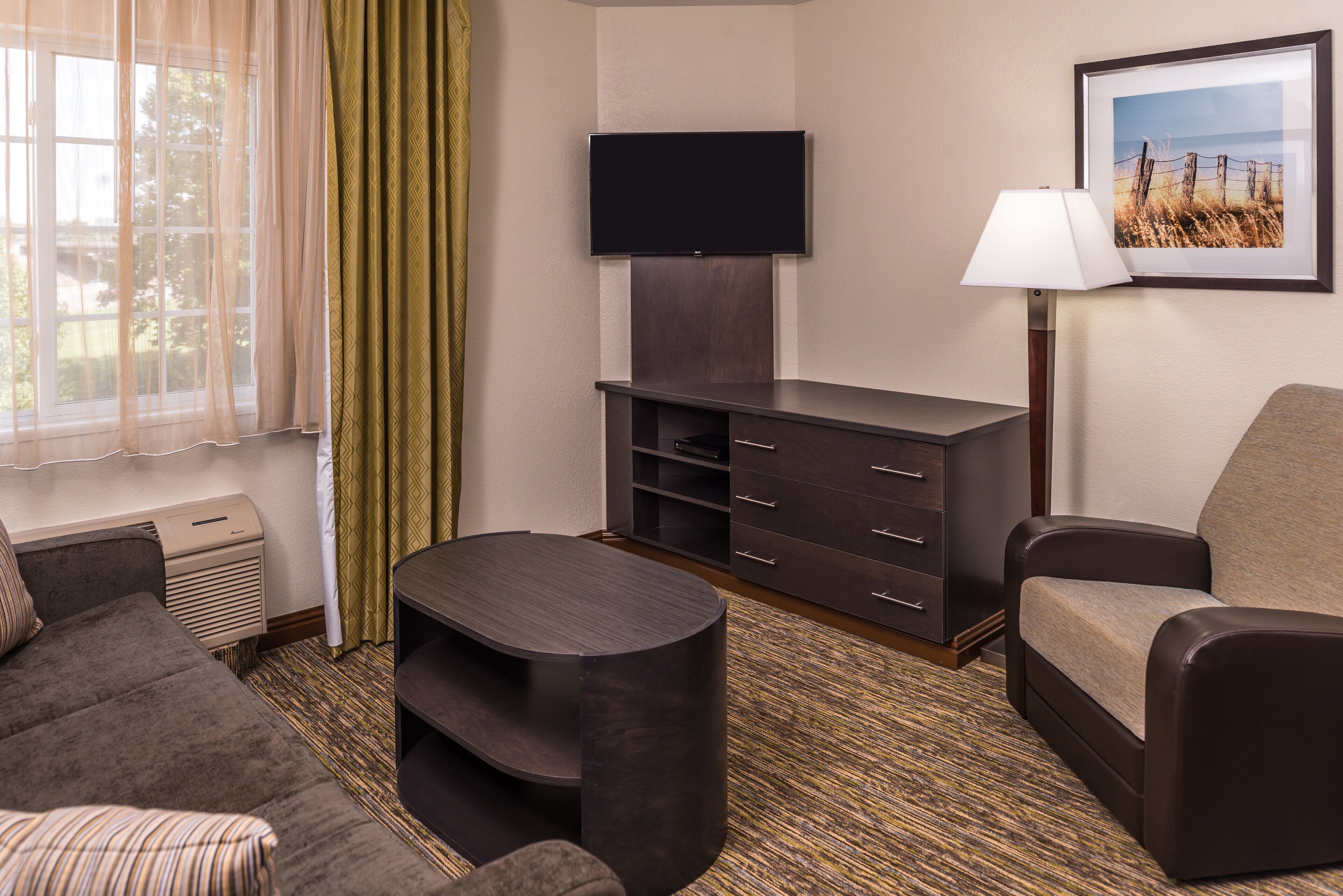 A hotel room with a flat-screen TV mounted on the wall, a sofa, a small table, a lamp, and a picture frame on the wall.