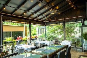 An outdoor patio area with tables and chairs, surrounded by greenery and large windows.