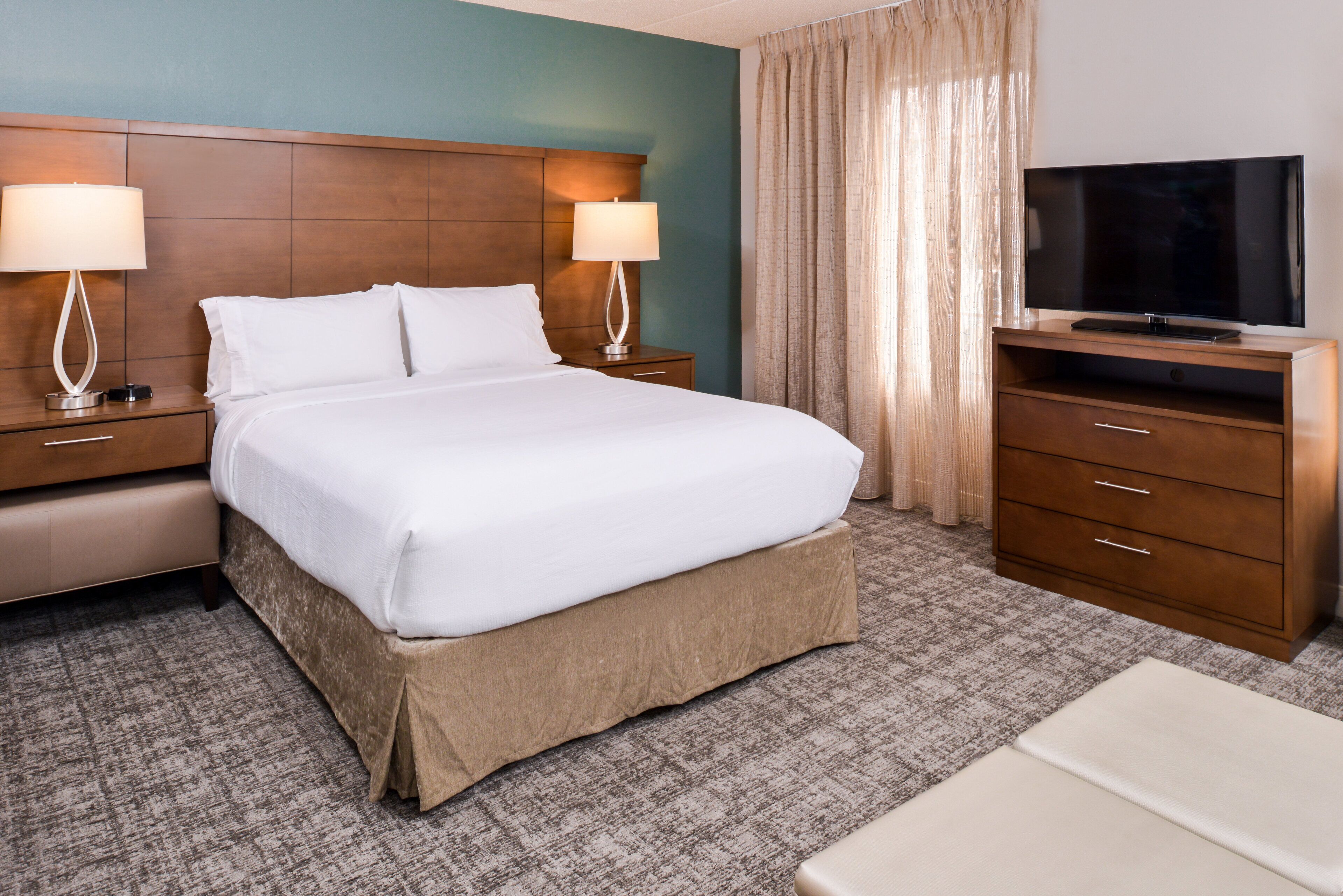 A hotel room with a large bed, two bedside tables with lamps, a TV on a wooden stand, and a carpeted floor.