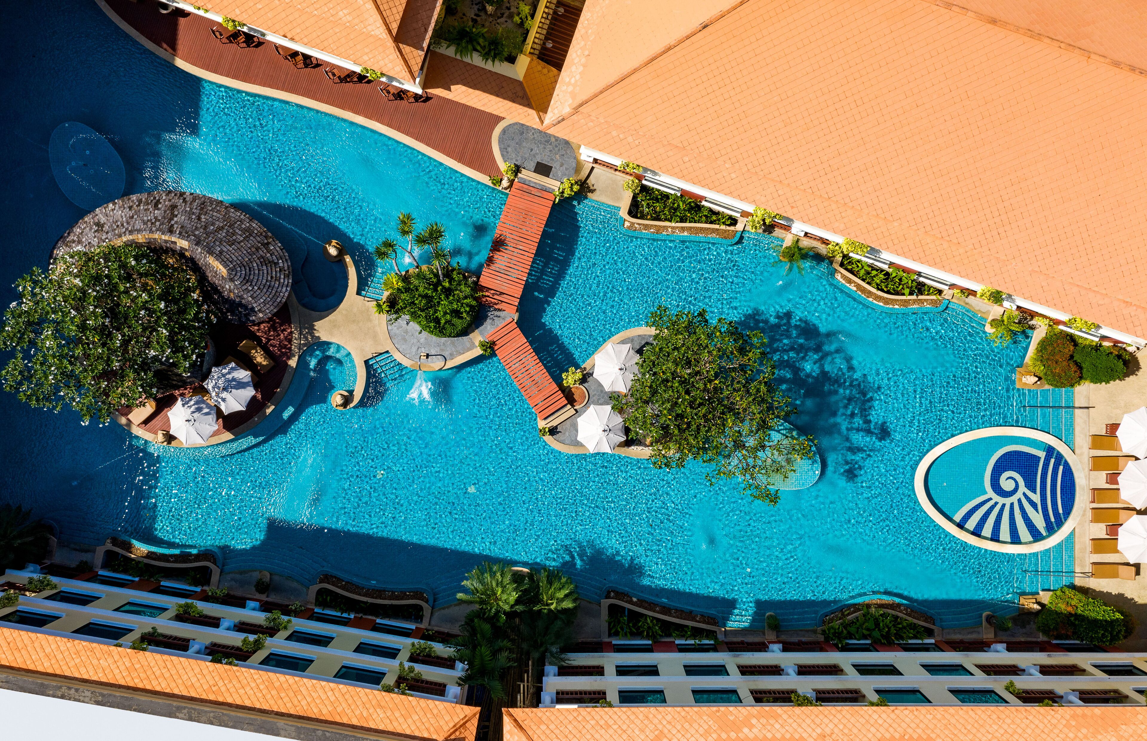 An aerial view of a resort pool area with multiple seating areas, a central water feature, and surrounding greenery.
