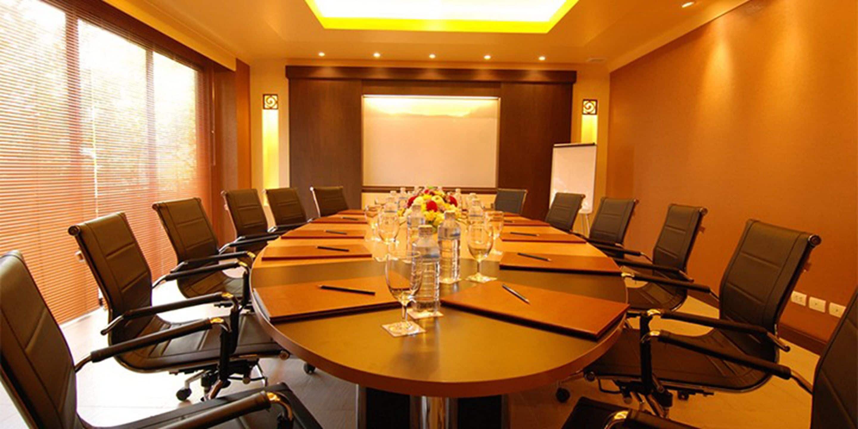 A conference room with a large oval table, multiple chairs, and a projector screen.