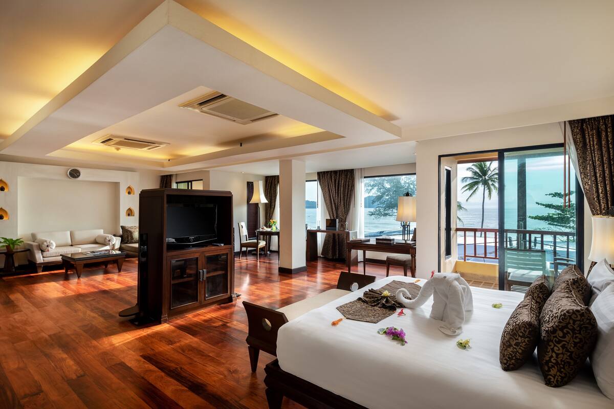 A hotel room with a wooden floor, a bed with a swan-shaped pillow, a TV, a desk, and a balcony with a view of the ocean.
