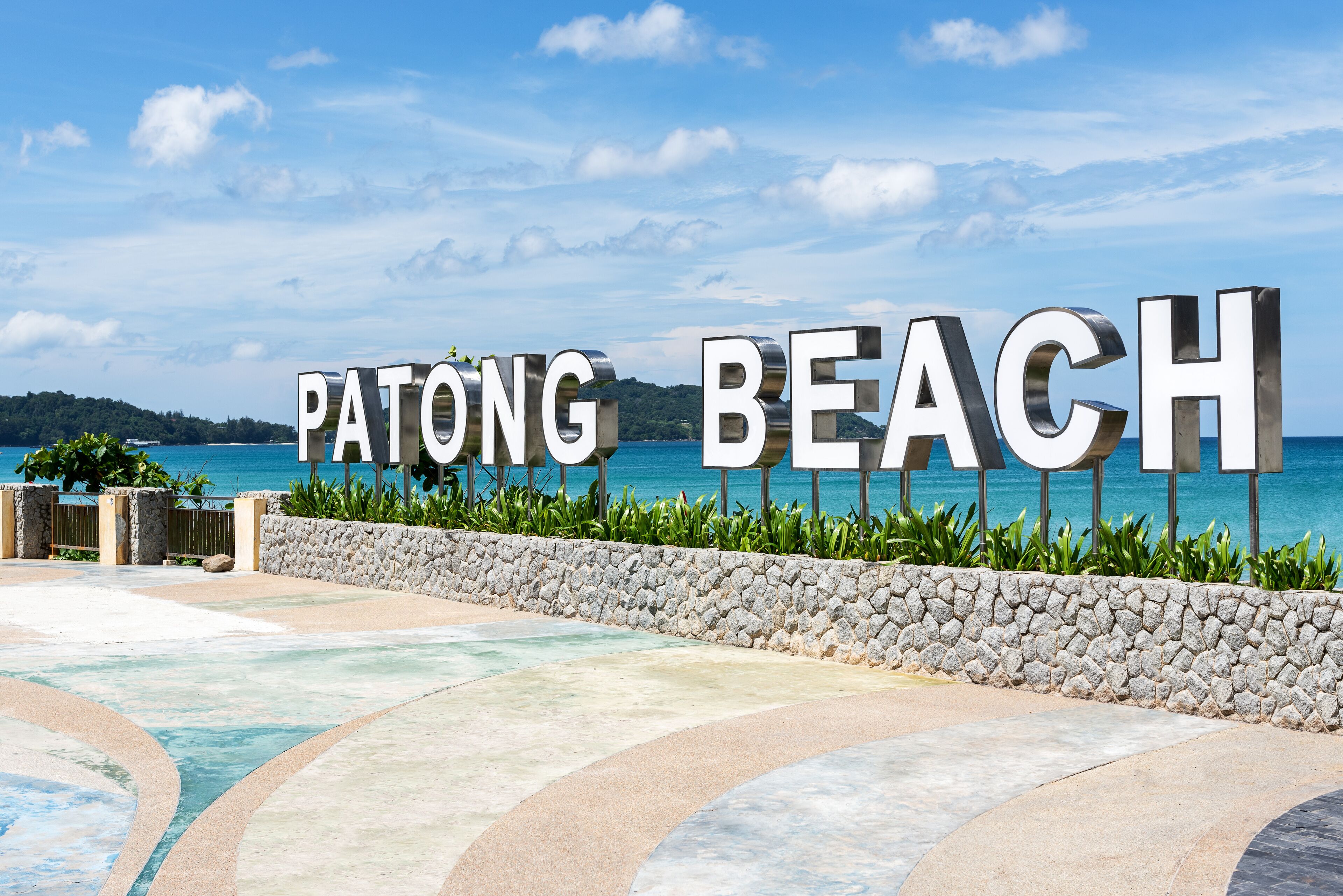 A sign reading "PATONG BEACH" with a view of the ocean and a stone wall.