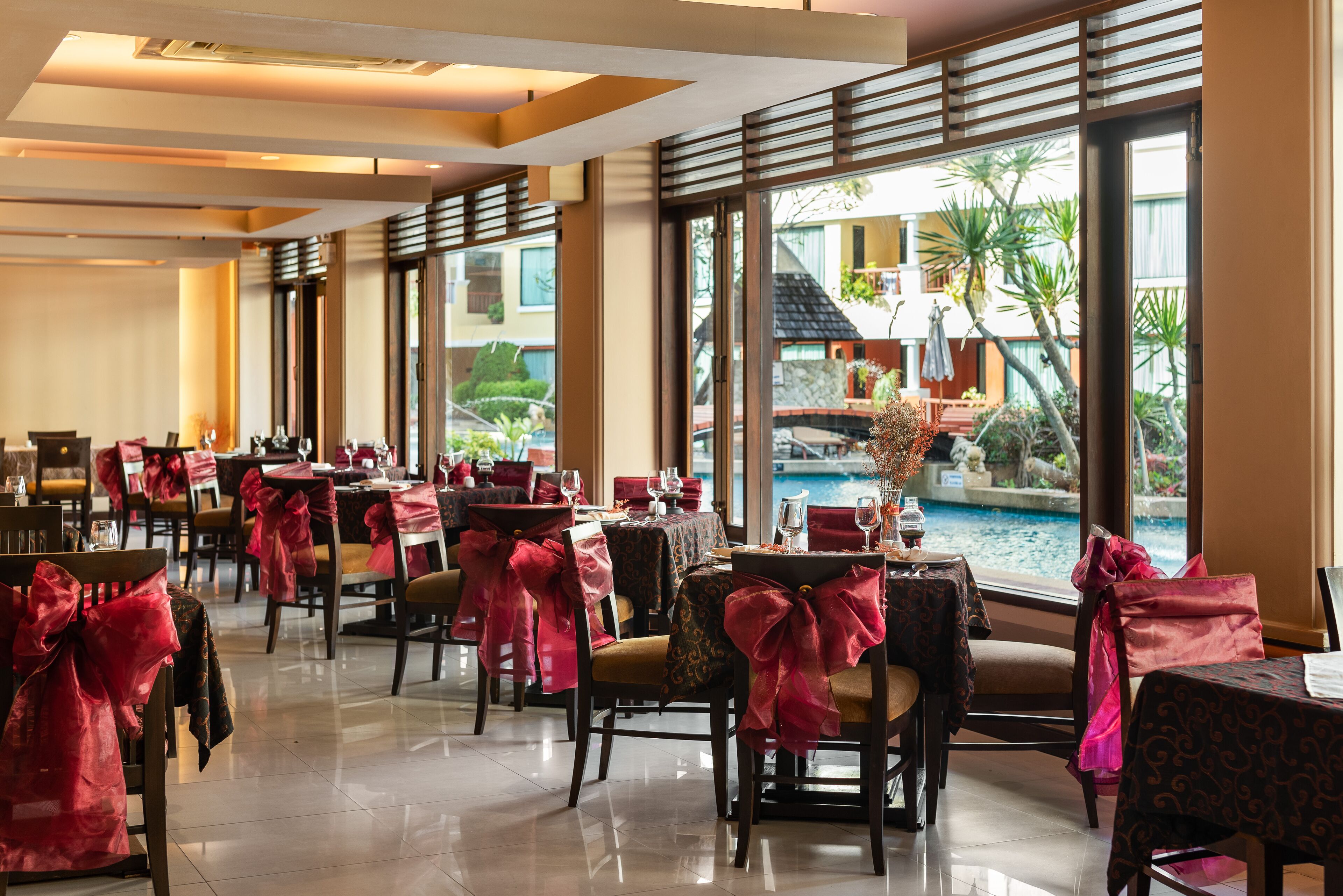 A restaurant with tables and chairs, large windows, and a view of a pool and greenery.