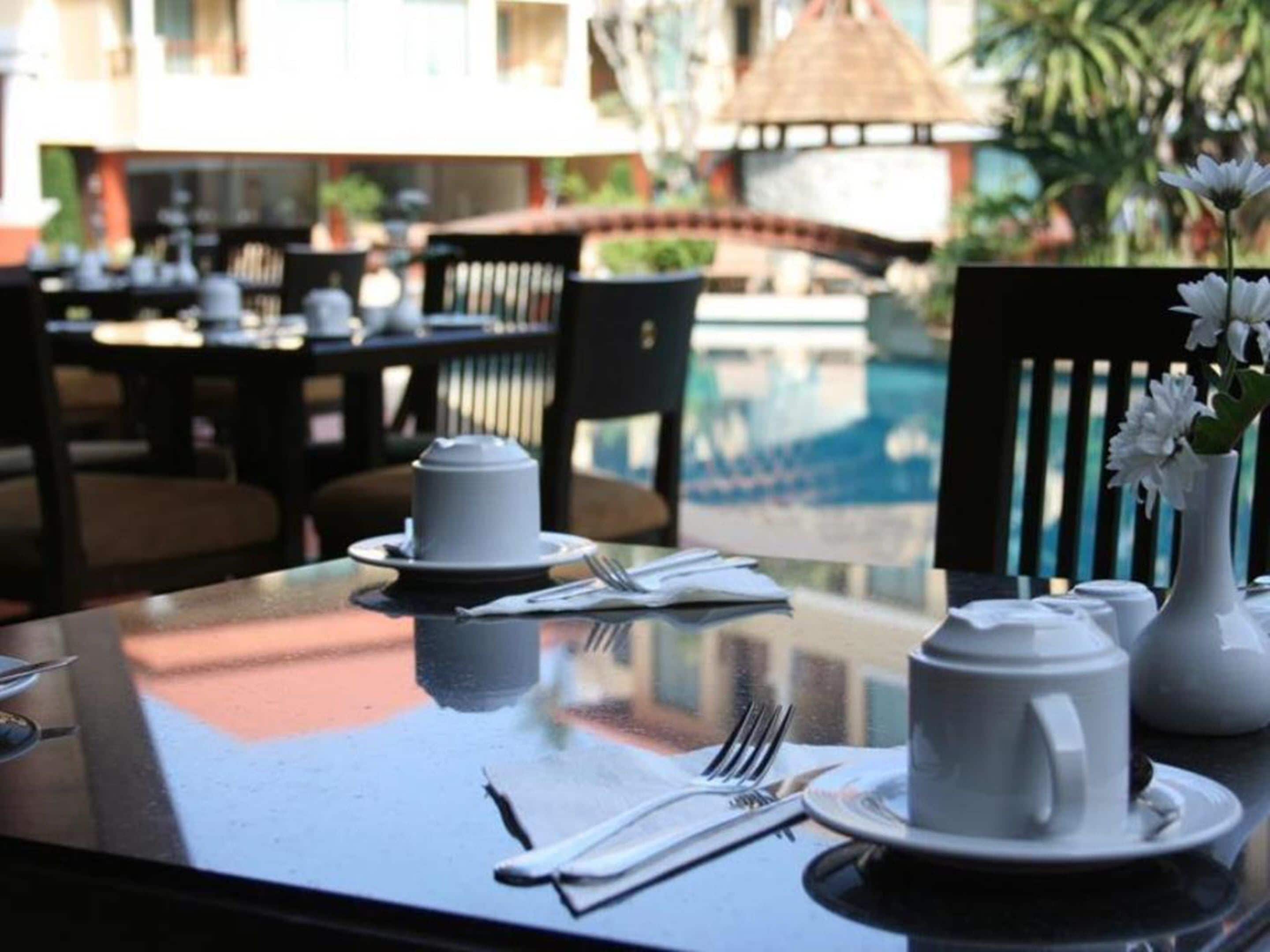 A dining table set with white dishes, a vase with flowers, and a view of an outdoor area with tables and chairs.