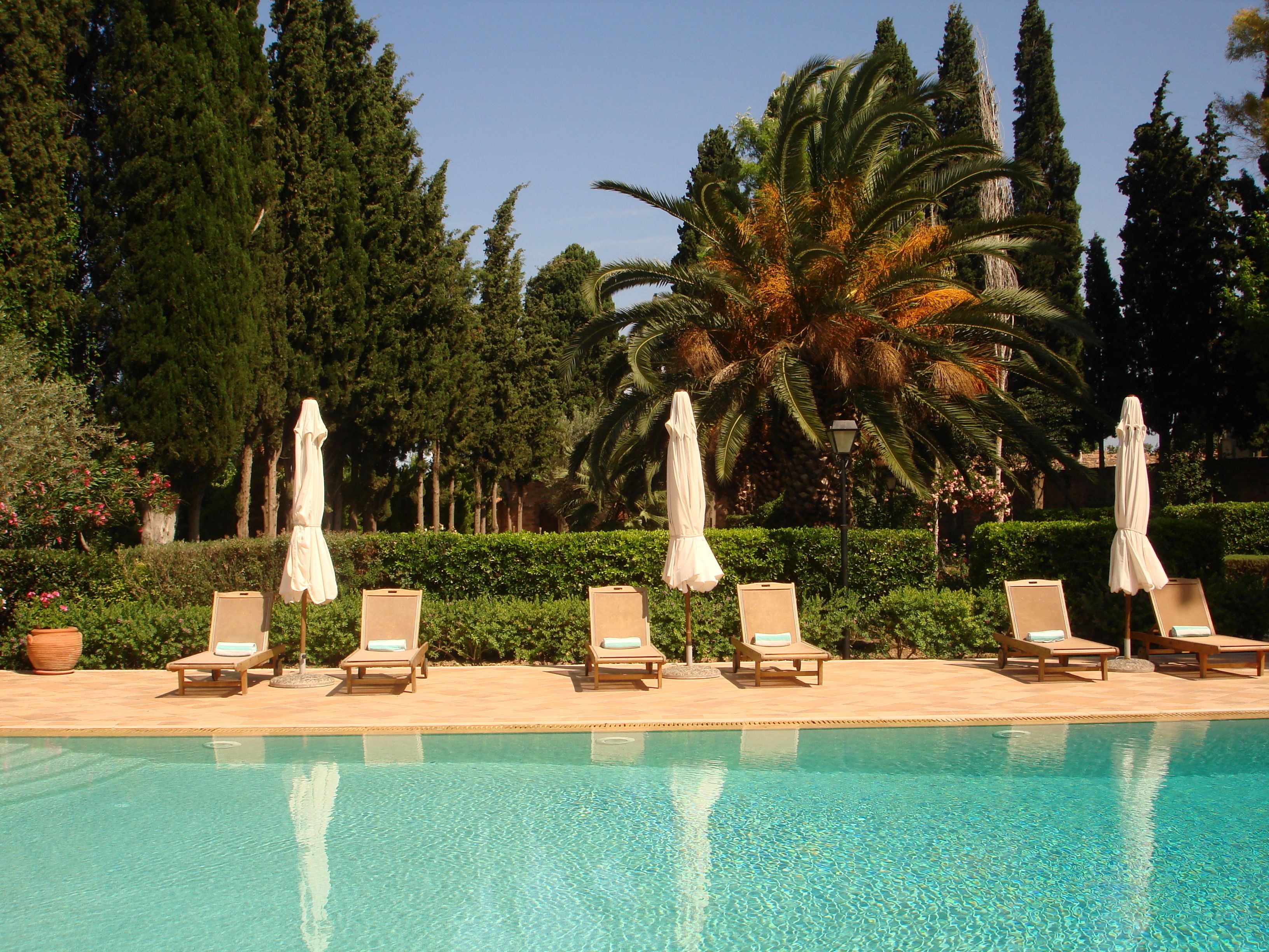 A poolside area with lounge chairs and umbrellas, surrounded by greenery and tall trees.