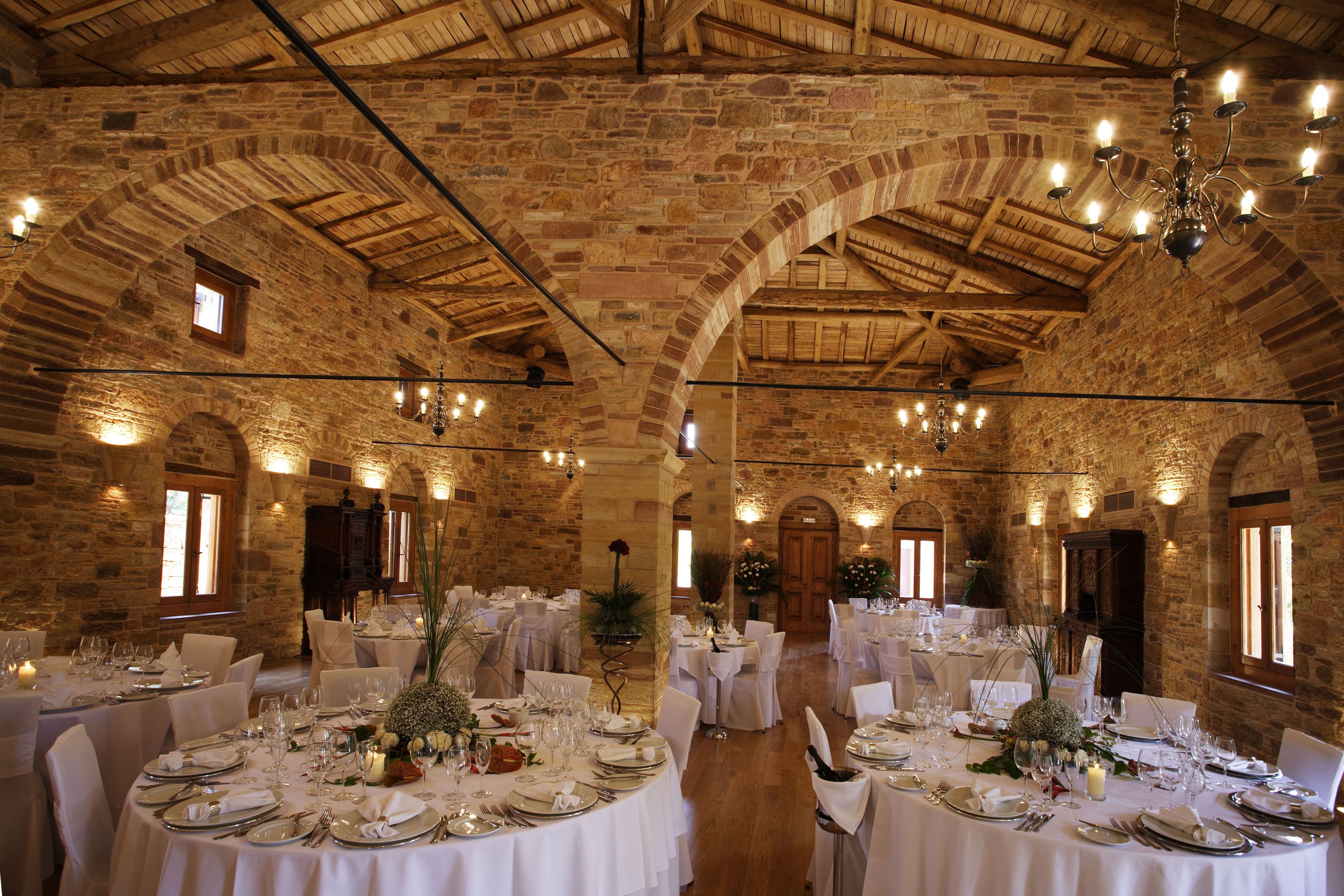 A spacious hall with stone walls and wooden beams, furnished with round tables set for dining, and chandeliers hanging from the ceiling.