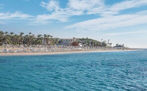 Un resort sulla spiaggia con file di lettini e ombrelloni, palme e un grande edificio sullo sfondo.