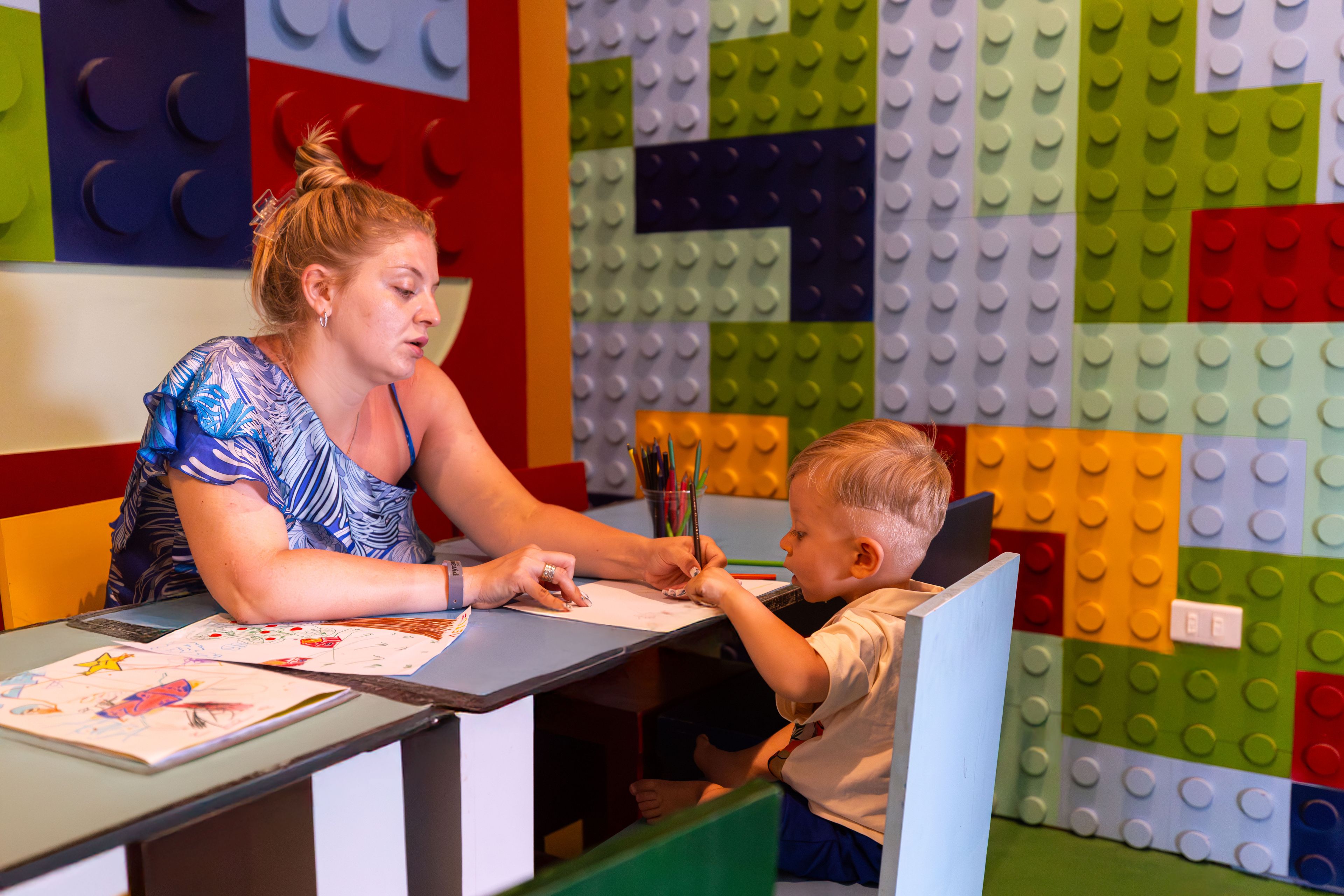 Una mujer y un niño en una mesa con papeles y un bolígrafo, en una habitación con paredes de LEGO de colores.