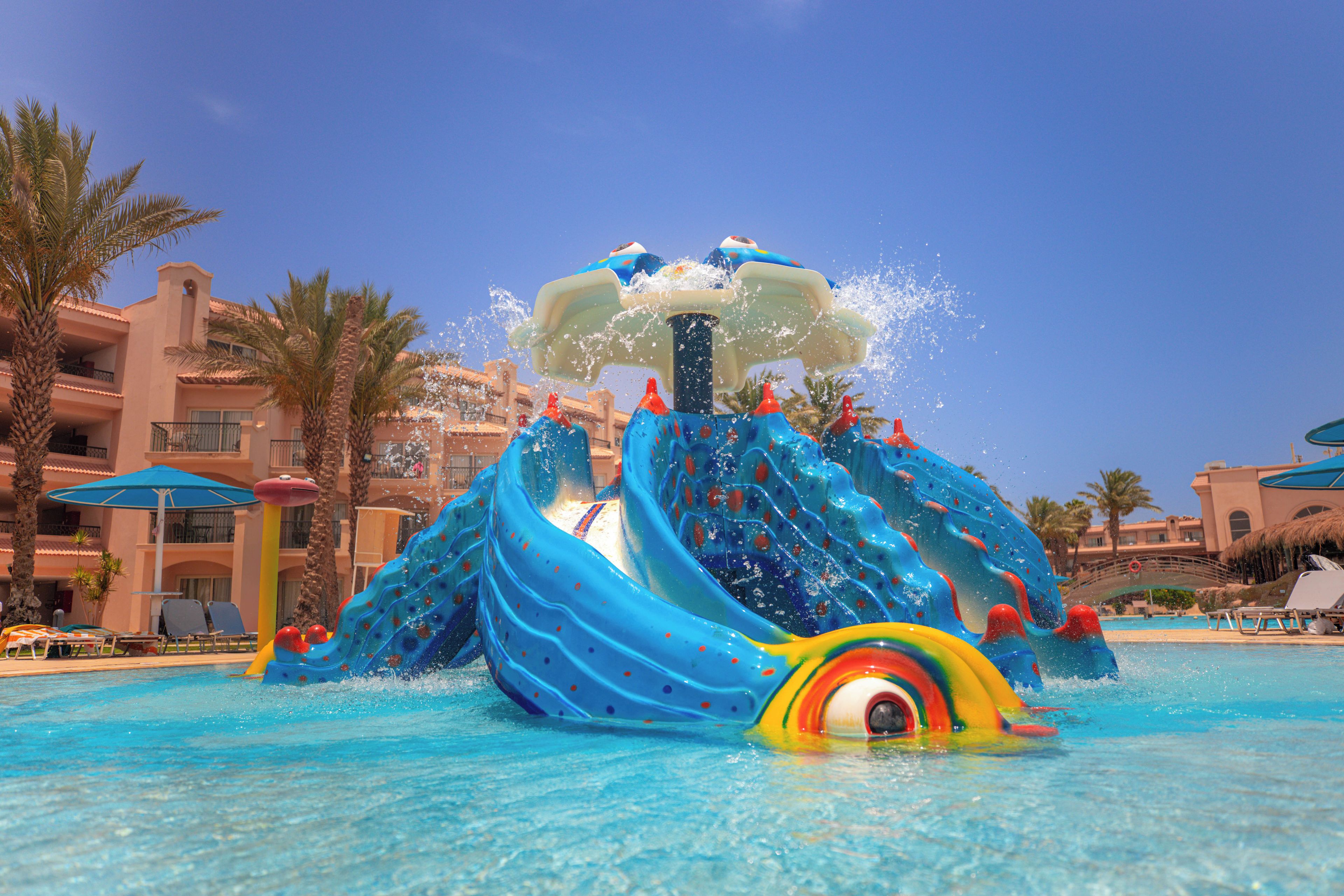 A water slide with a dolphin theme, surrounded by a pool, palm trees, and hotel buildings.