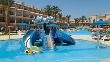 Relax by the pool surrounded by lounge chairs and umbrellas, with a central water slide and Mediterranean-style resort buildings in the background.