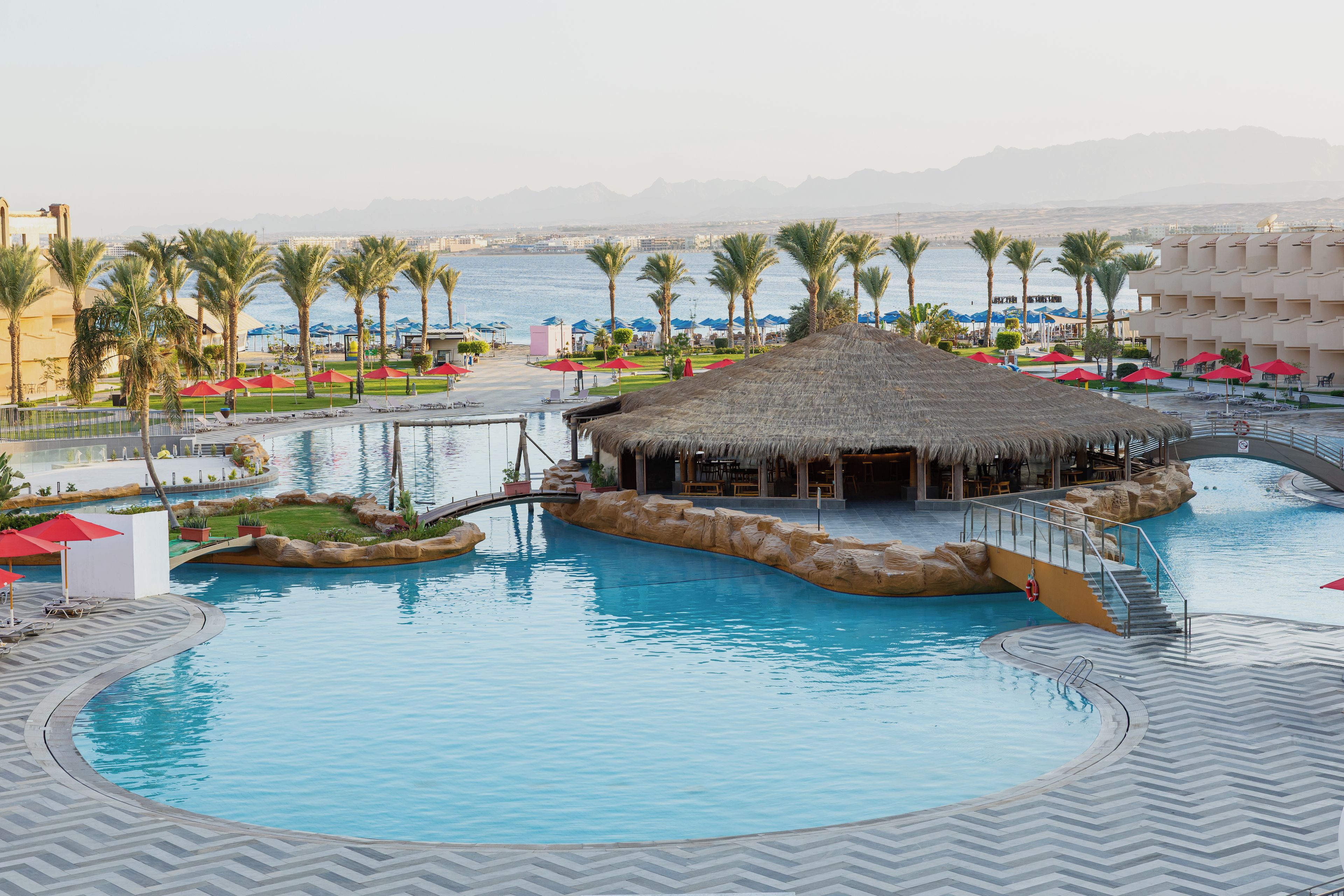 A resort pool area with a central water feature, palm trees, and a thatched-roof structure.