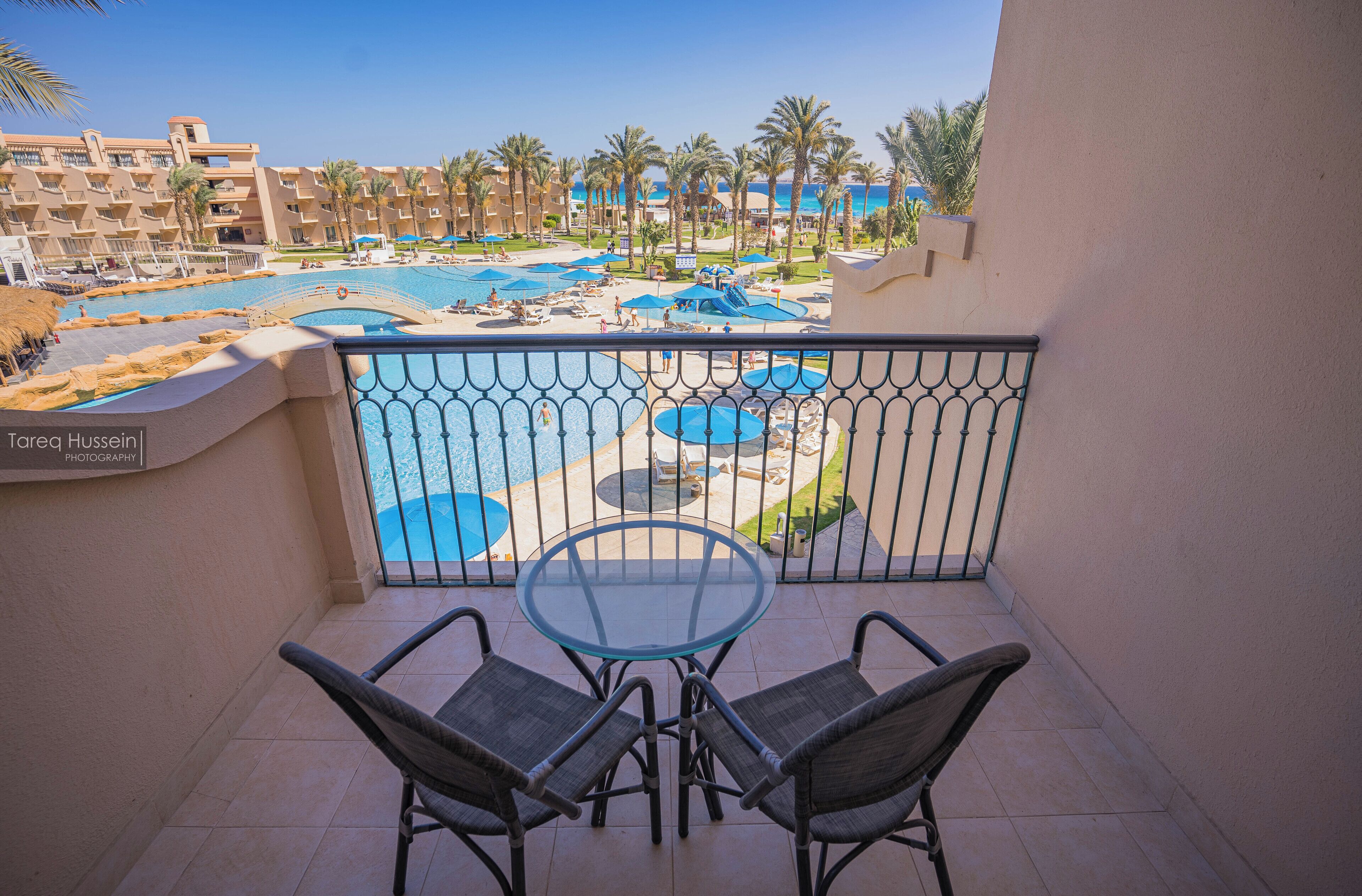 A balcony with a glass table and two chairs, overlooking a pool area with multiple pools and palm trees.