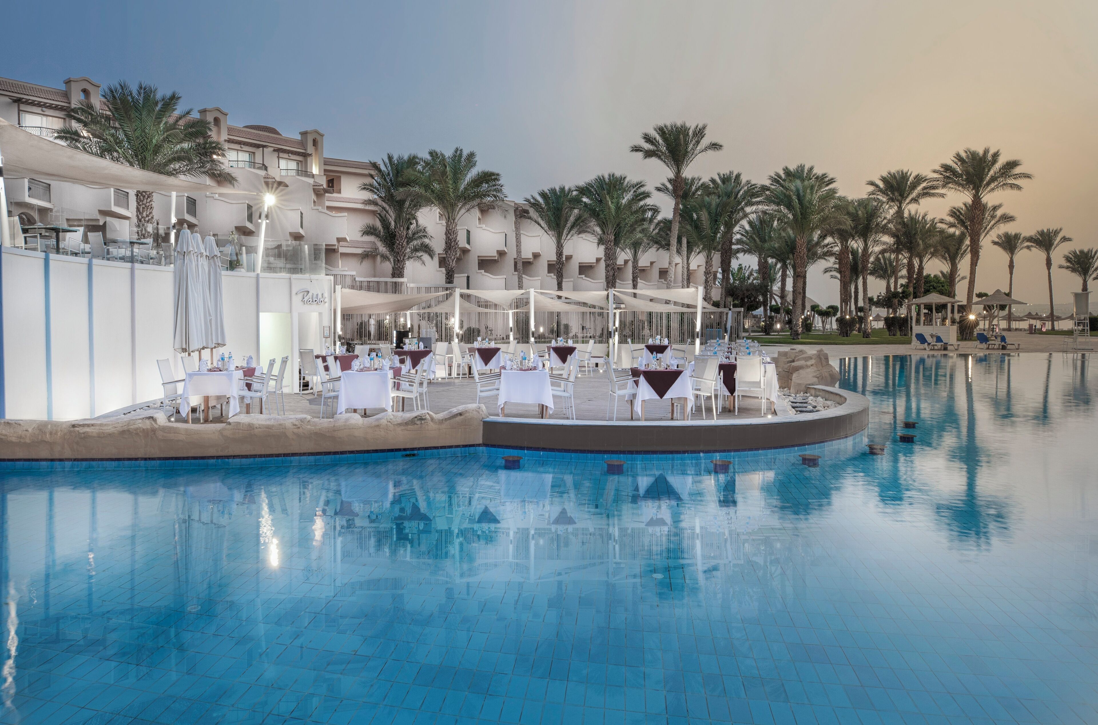 Área de piscina de resort com piscina circular, espreguiçadeiras brancas e mesas montadas para refeições. Palmeiras e um prédio com varandas são visíveis ao fundo.