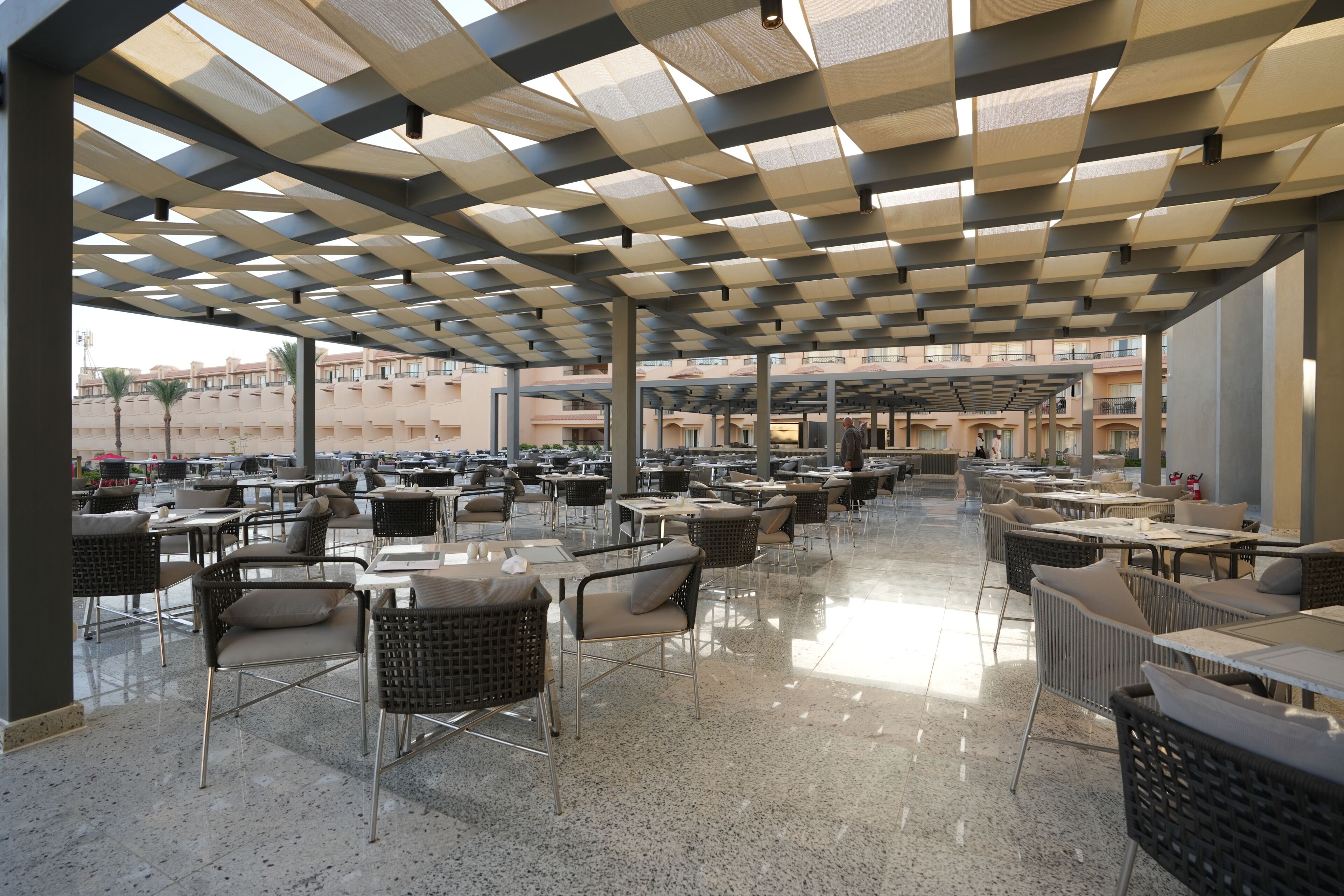 An outdoor cafe area with multiple tables and chairs, a checkered ceiling, and a view of buildings in the background.