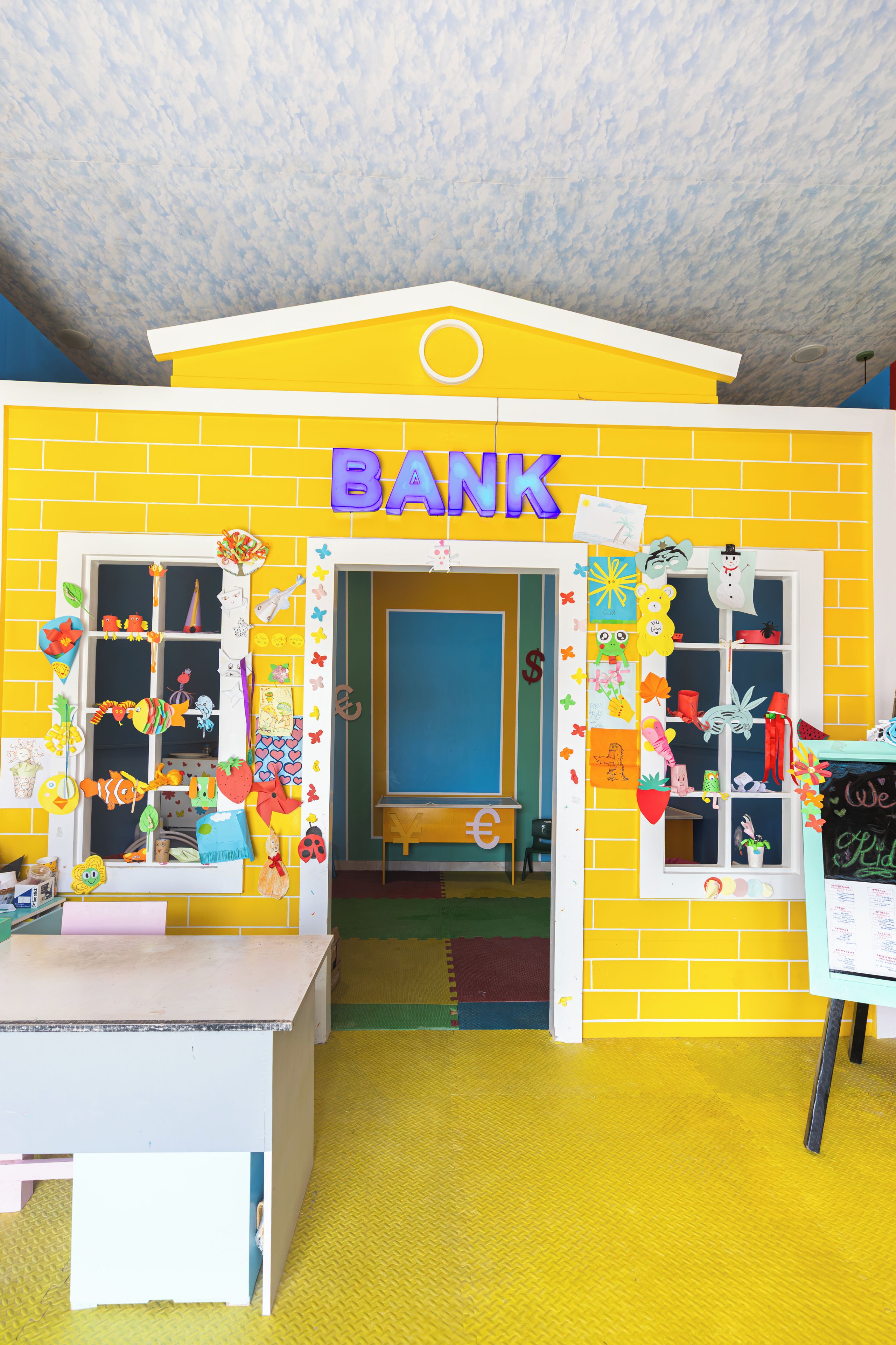 Een kleurrijke binnenspeelruimte, ingericht als een bank met een speeltafel en een prikbordwand.