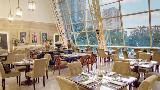 Un restaurante amplio con techos altos y ventanas grandes que ofrecen vistas de la ciudad. Las mesas están ordenadas con sillas, y el interior presenta una decoración e iluminación modernos.