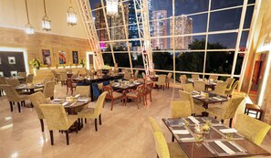 A restaurant with multiple dining tables and chairs, large windows offering a city view, and decorative lighting.