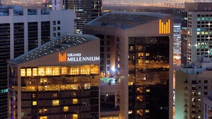 Un hotel de varios pisos con un llamativo letrero de 'Grand Millennium', que presenta un diseño arquitectónico moderno con ventanas iluminadas y una terraza en la azotea.