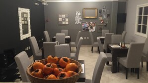 Zona pranzo con diversi tavoli e sedie, una parete con quadri e una bacheca, e un cesto di pomodori sul tavolo.