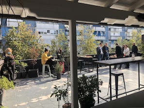 En udendørs terrasse med borde og stole, krukker med planter og udsigt til boligbygninger.