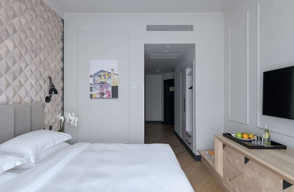 A hotel room with a bed, a desk with a flat-screen TV, a painting on the wall, and a door leading to another room.