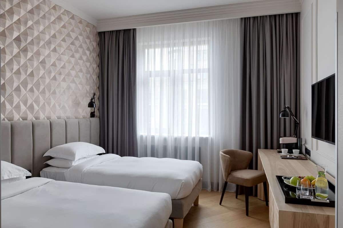 A hotel room with two beds, a desk with a chair, a TV, and a breakfast bar with fruit.