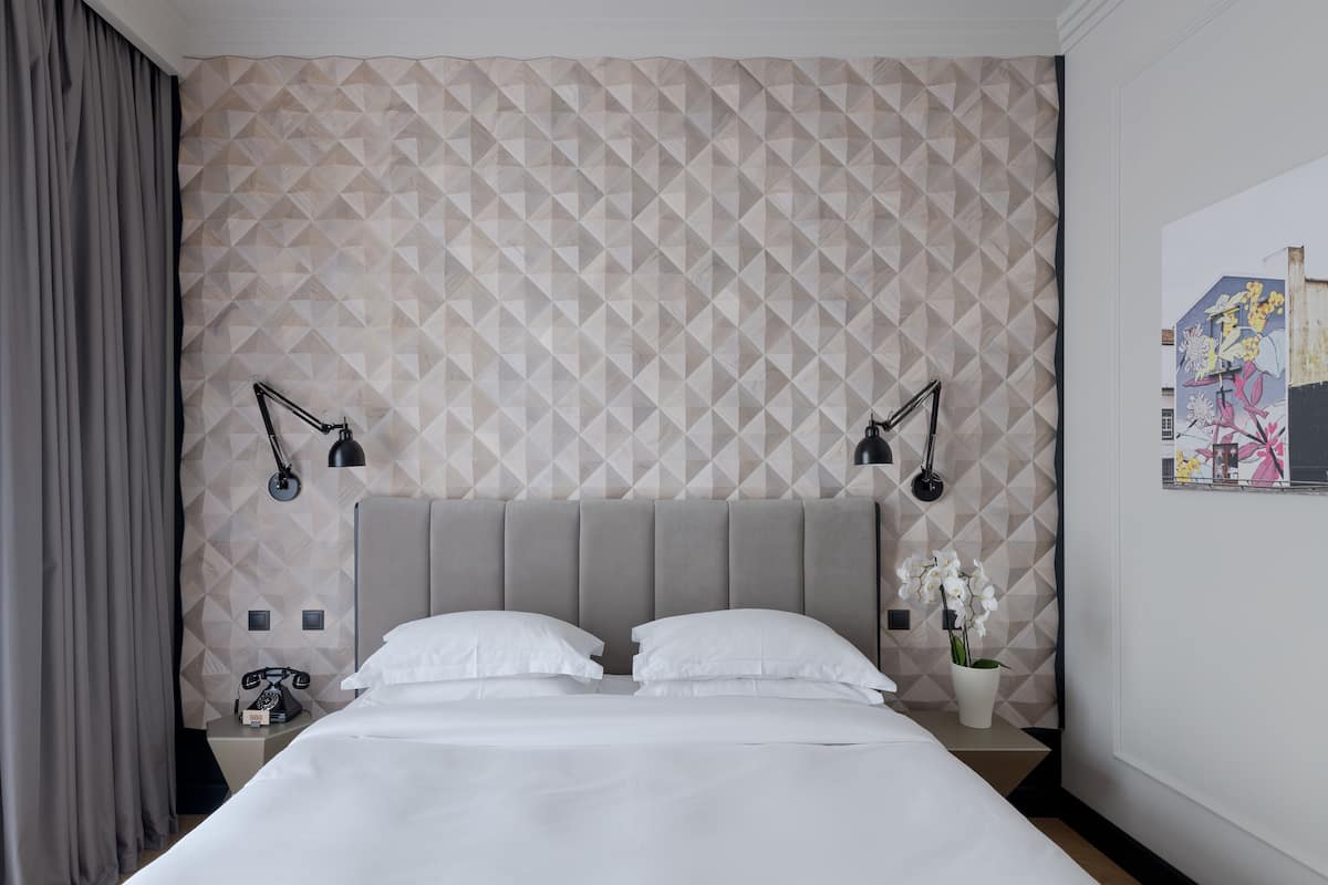 A hotel room with a geometric patterned wall, a bed with white linens, two bedside lamps, a telephone, and a small potted plant.