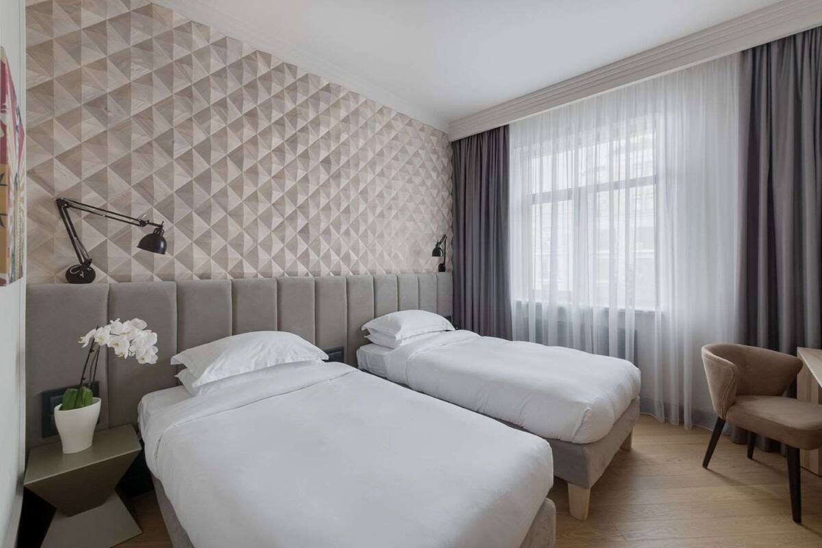 A hotel room with two beds, a desk, a chair, a lamp, and a geometric patterned wall.