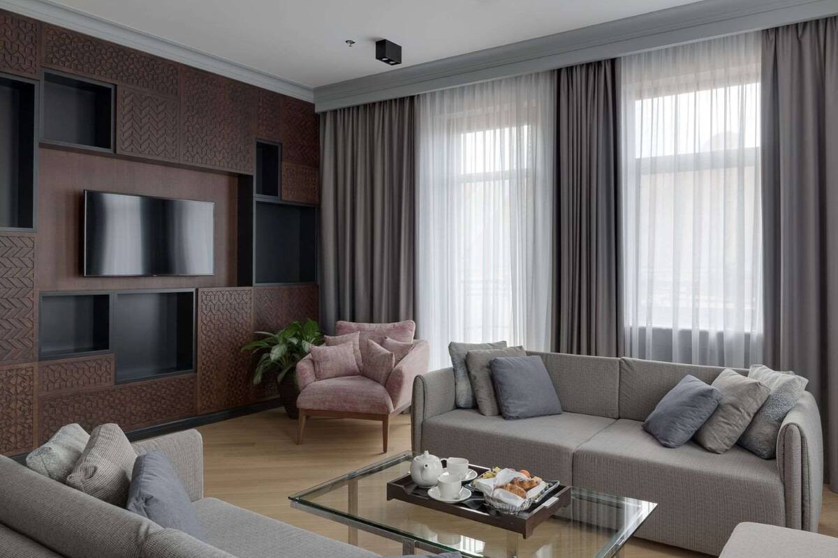 A modern living room with a flat-screen TV mounted on a textured wall, a grey sofa with cushions, a glass coffee table with a tray of breakfast items, and large windows with sheer curtains.