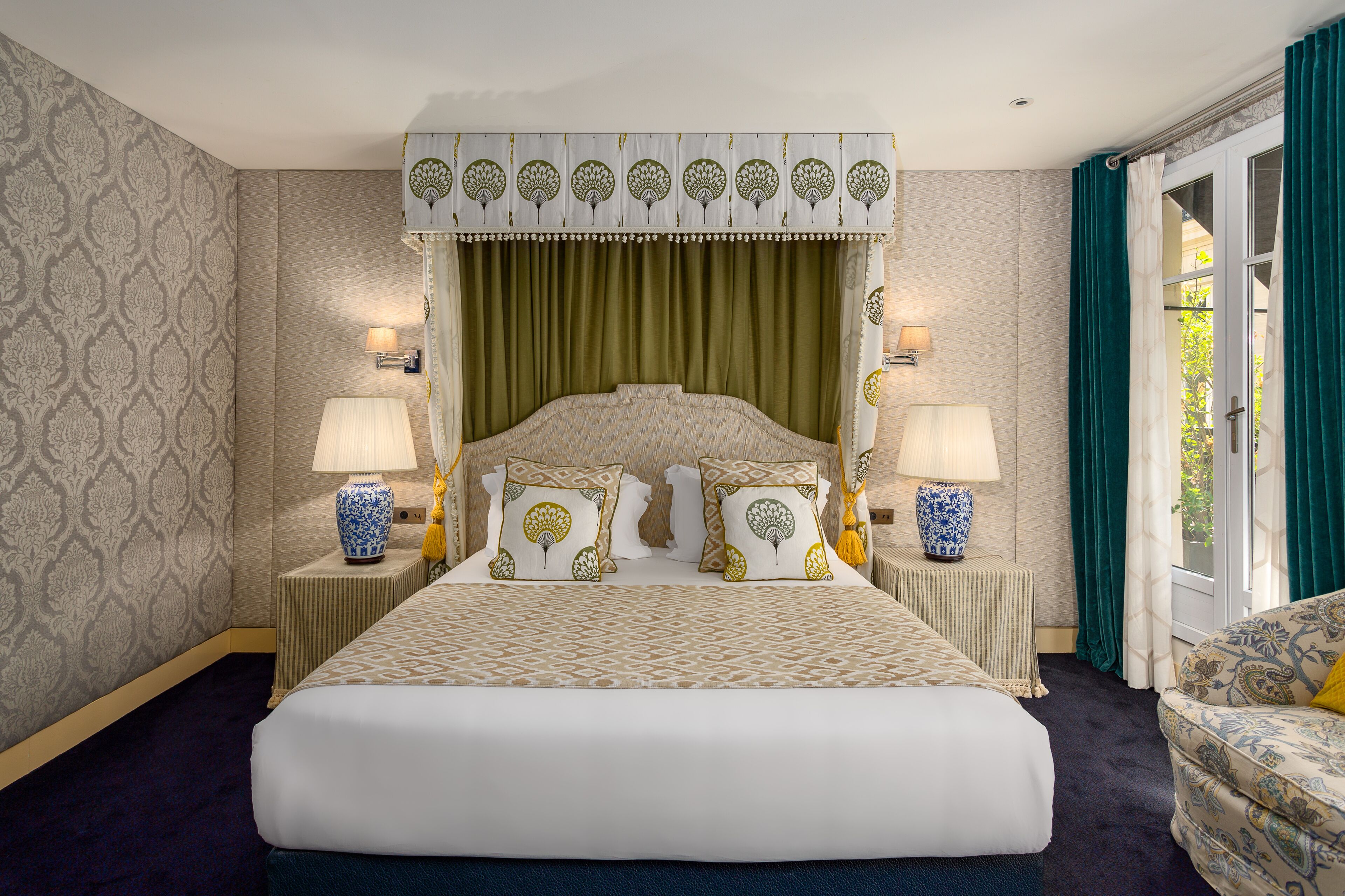 Une chambre d’hôtel avec un grand lit, deux tables de chevet avec des lampes, un papier peint à motifs et une vue sur l’extérieur, visible à travers une porte.