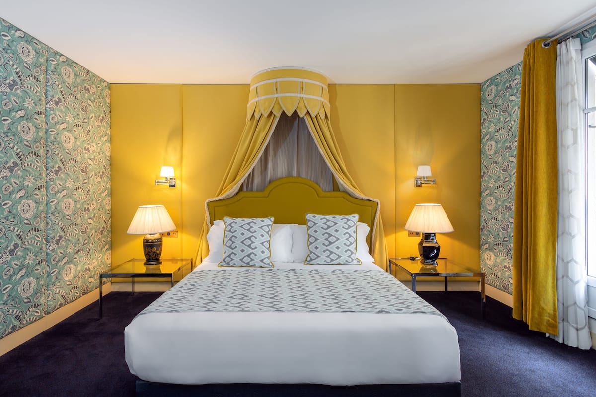 A hotel room with a large bed, two bedside tables with lamps, patterned wallpaper, and a yellow accent wall.