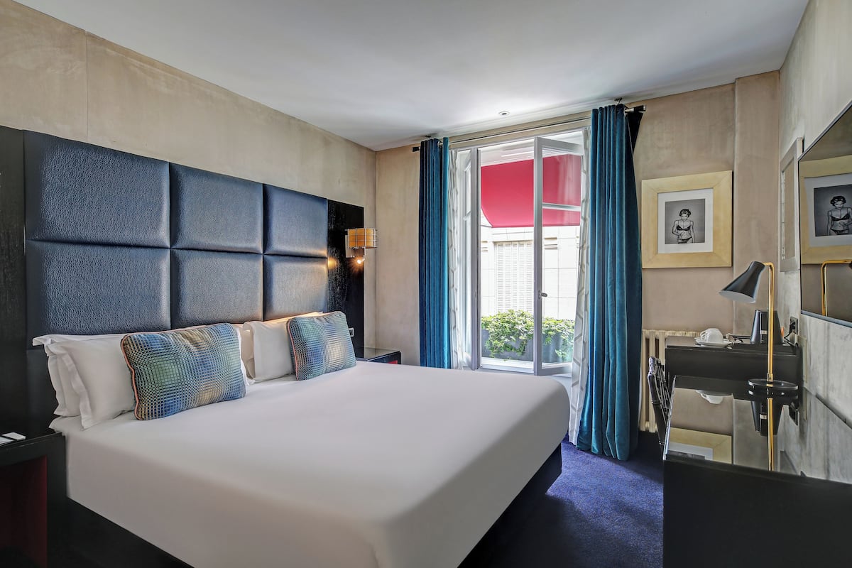 A hotel room with a large bed, a desk with a chair, a window with blue curtains, and a view of the outdoors.