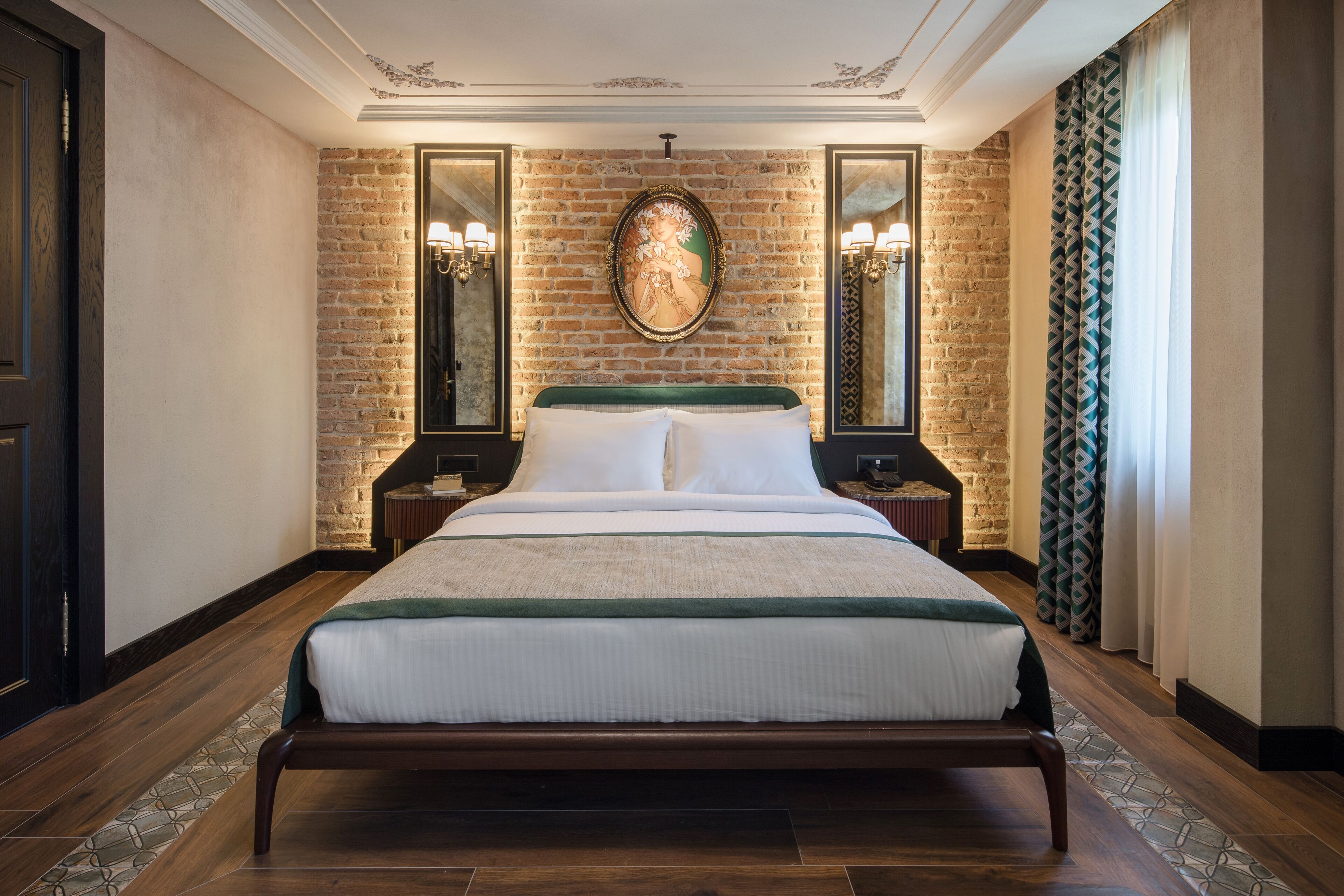 A large bed with a wooden frame, flanked by two nightstands with lamps, in a room with exposed brick walls and a large mirror above the bed.