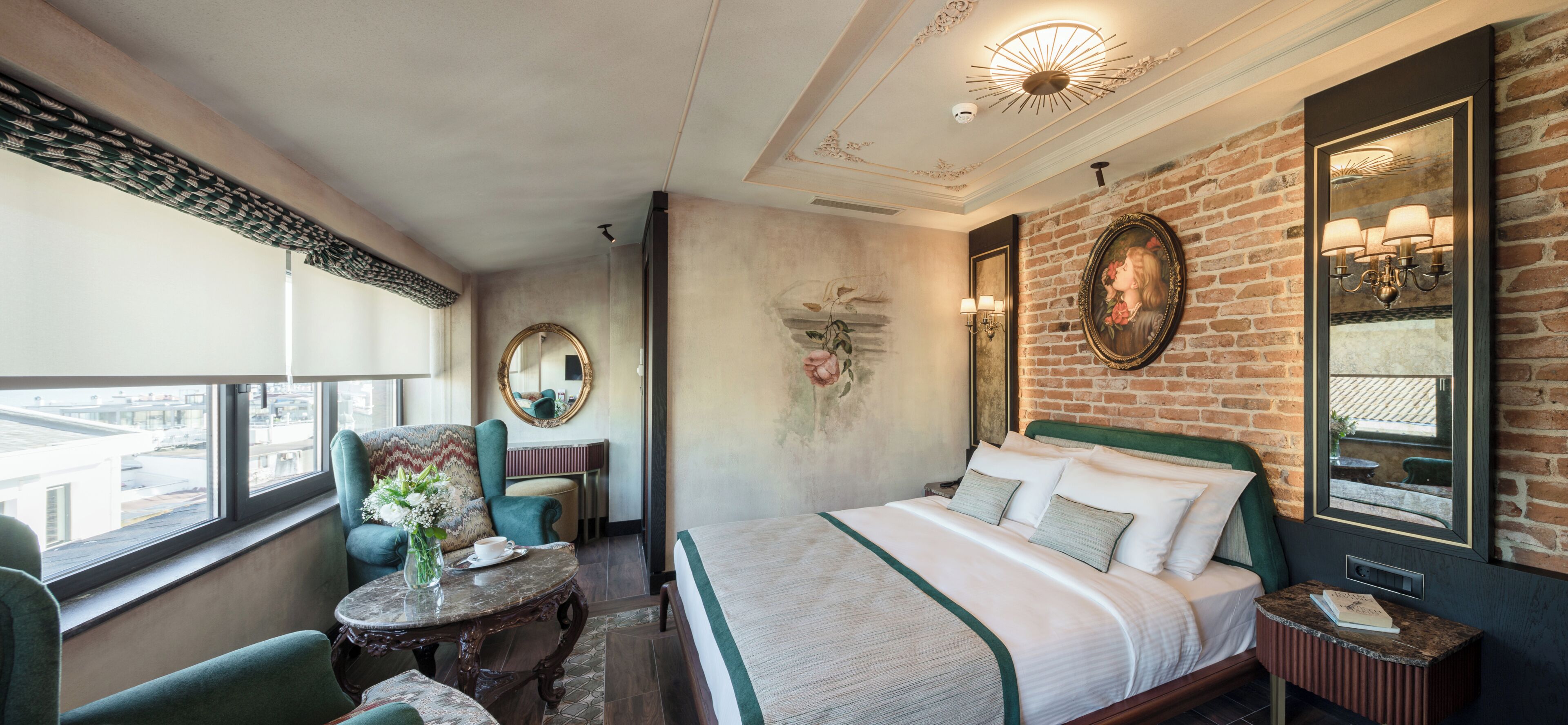 A hotel room with a large bed, a sitting area with a chair and table, a view through the window, and a brick wall.