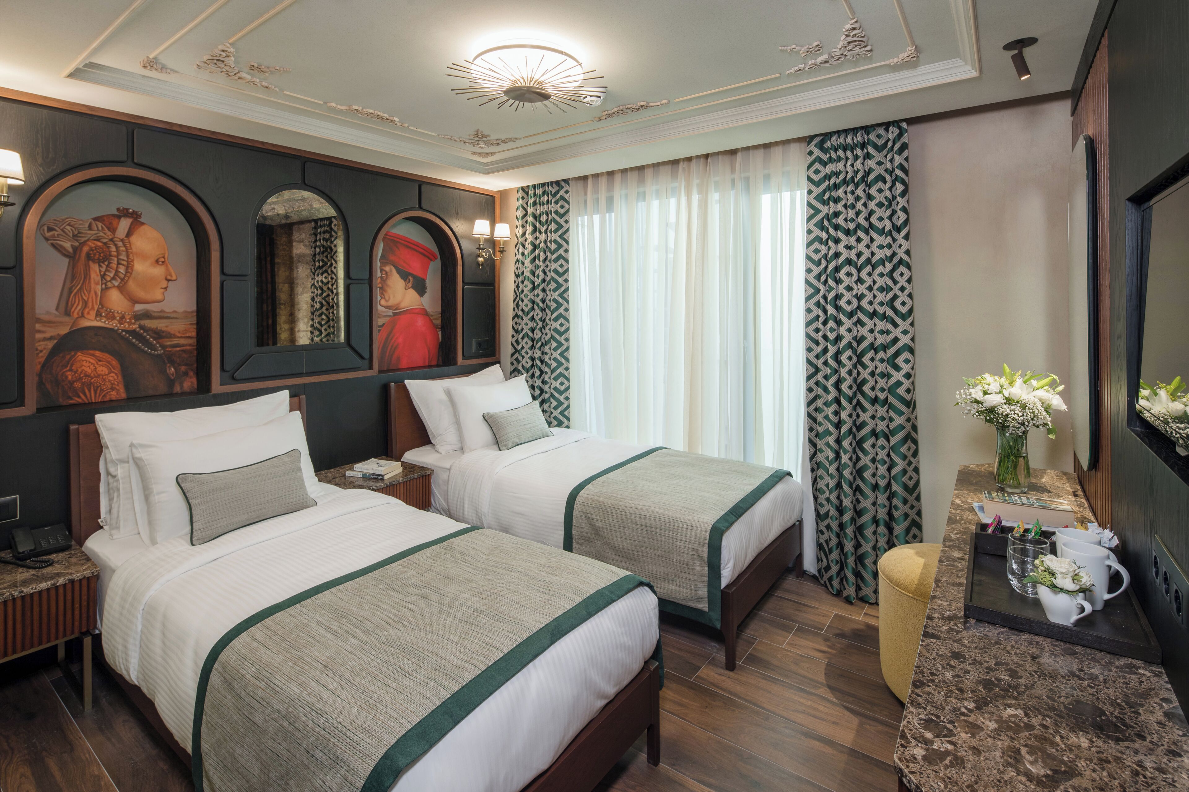 A hotel room with two beds, a nightstand, a desk with a lamp, a mirror, and a window with patterned curtains.