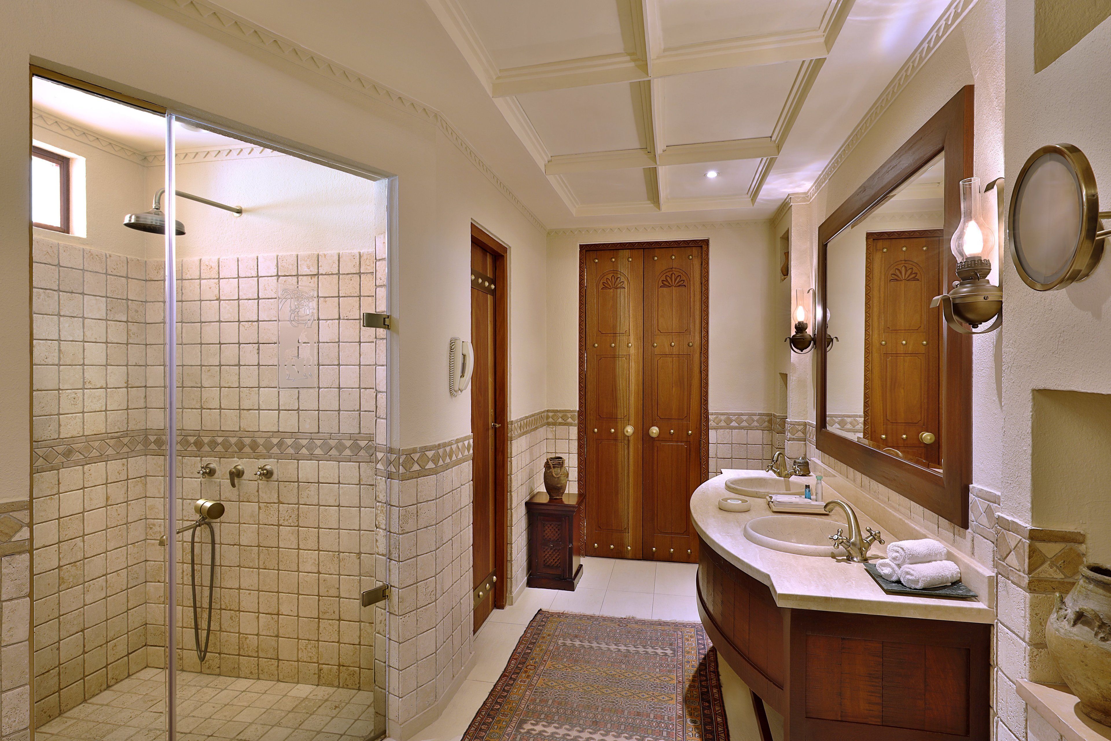 A bathroom with a glass shower enclosure, a sink with a mirror, and a patterned rug on the floor.