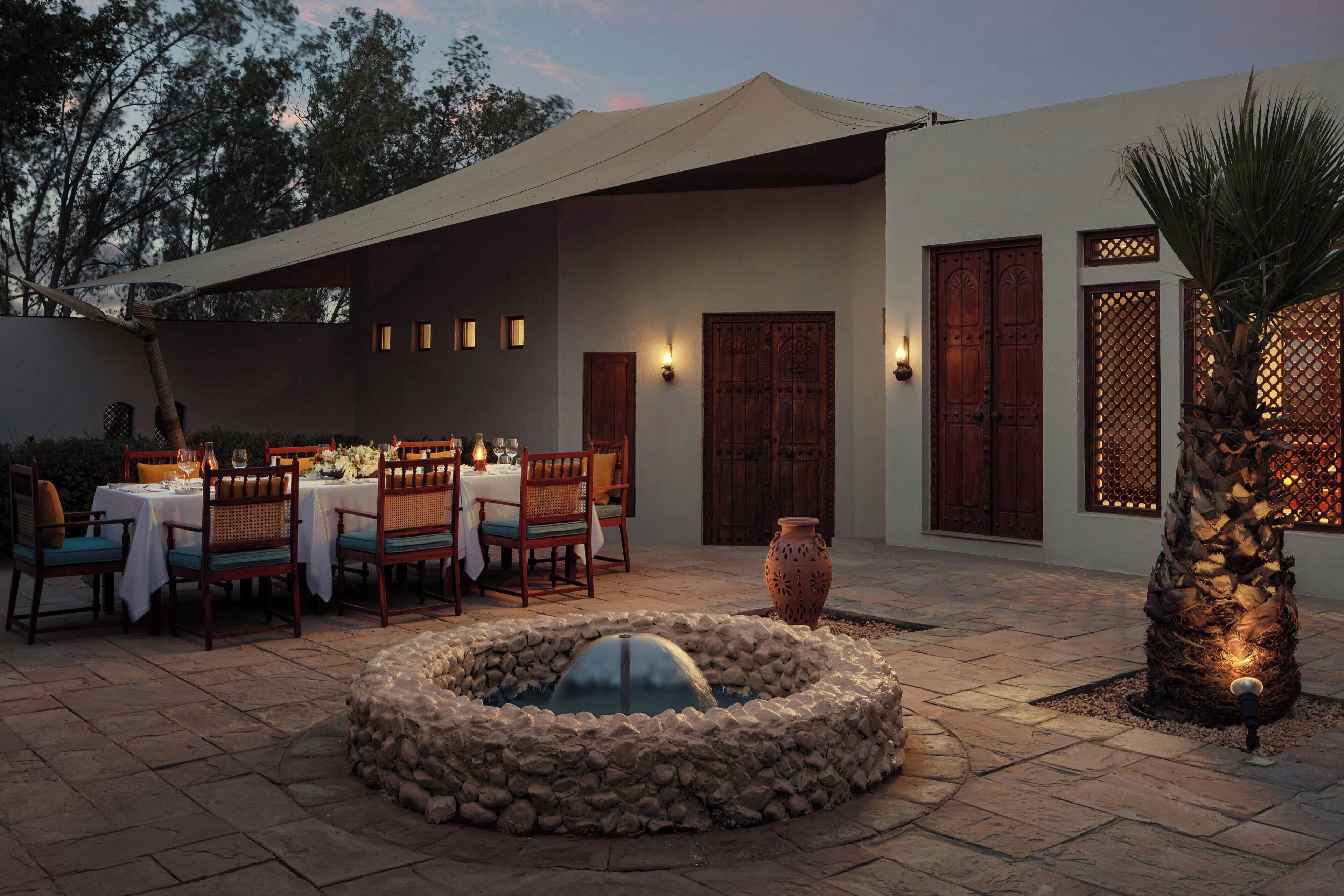 An outdoor dining area with tables and chairs, a stone fire pit, and a decorative palm tree.