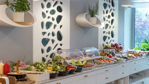 Un bufé con diversos alimentos, como ensaladas, frutas y sándwiches, dispuesto sobre una mesa blanca con paneles decorativos y plantas en macetas.