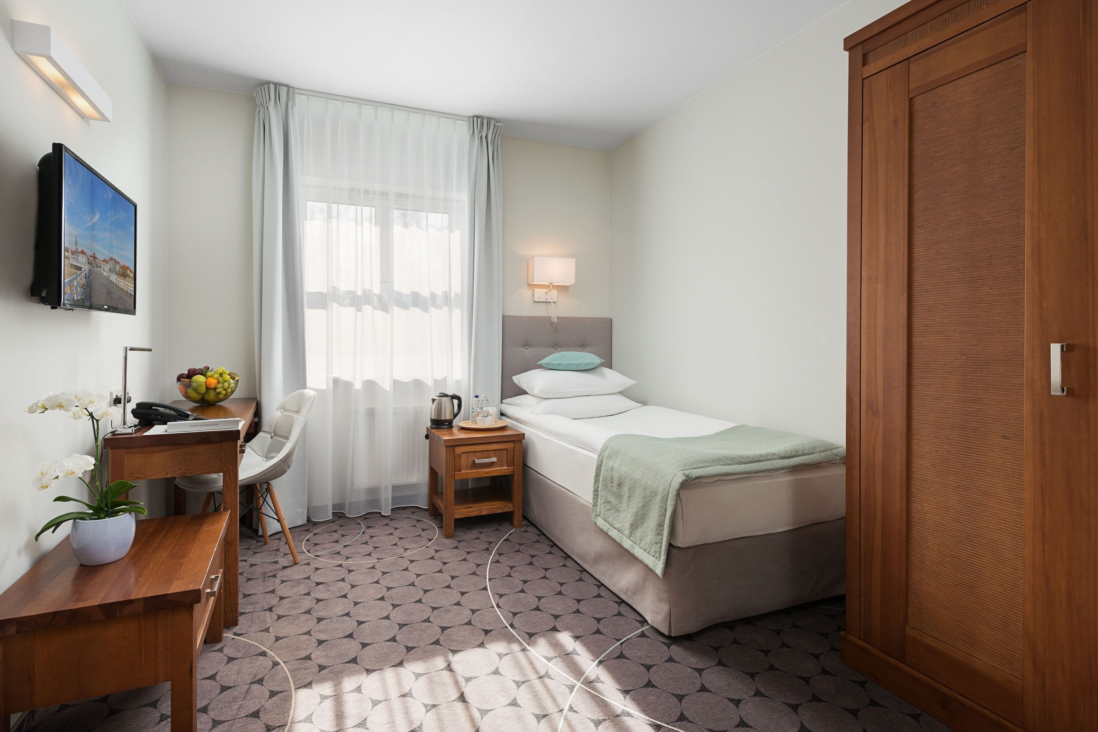 A hotel room with a bed, a desk, a chair, a nightstand, a lamp, a wardrobe, and a window with curtains.