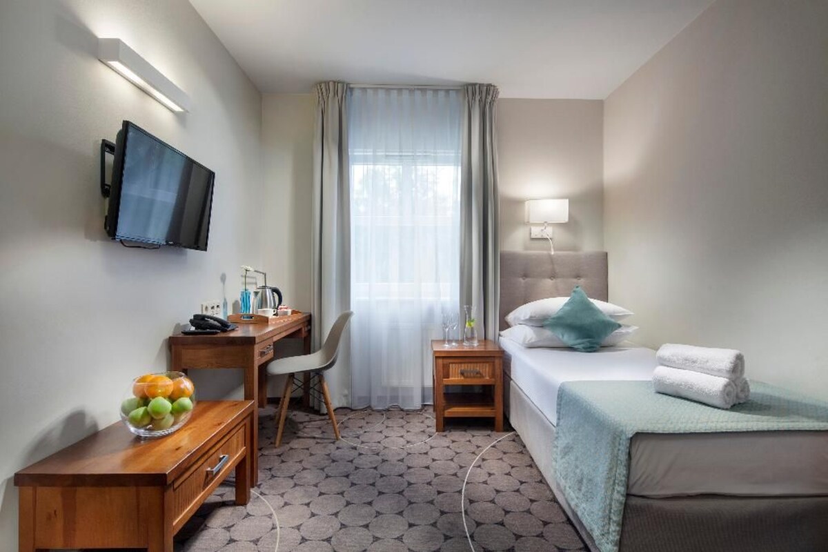 A hotel room with a bed, a desk with a chair, a television mounted on the wall, a window with curtains, and a bedside lamp.
