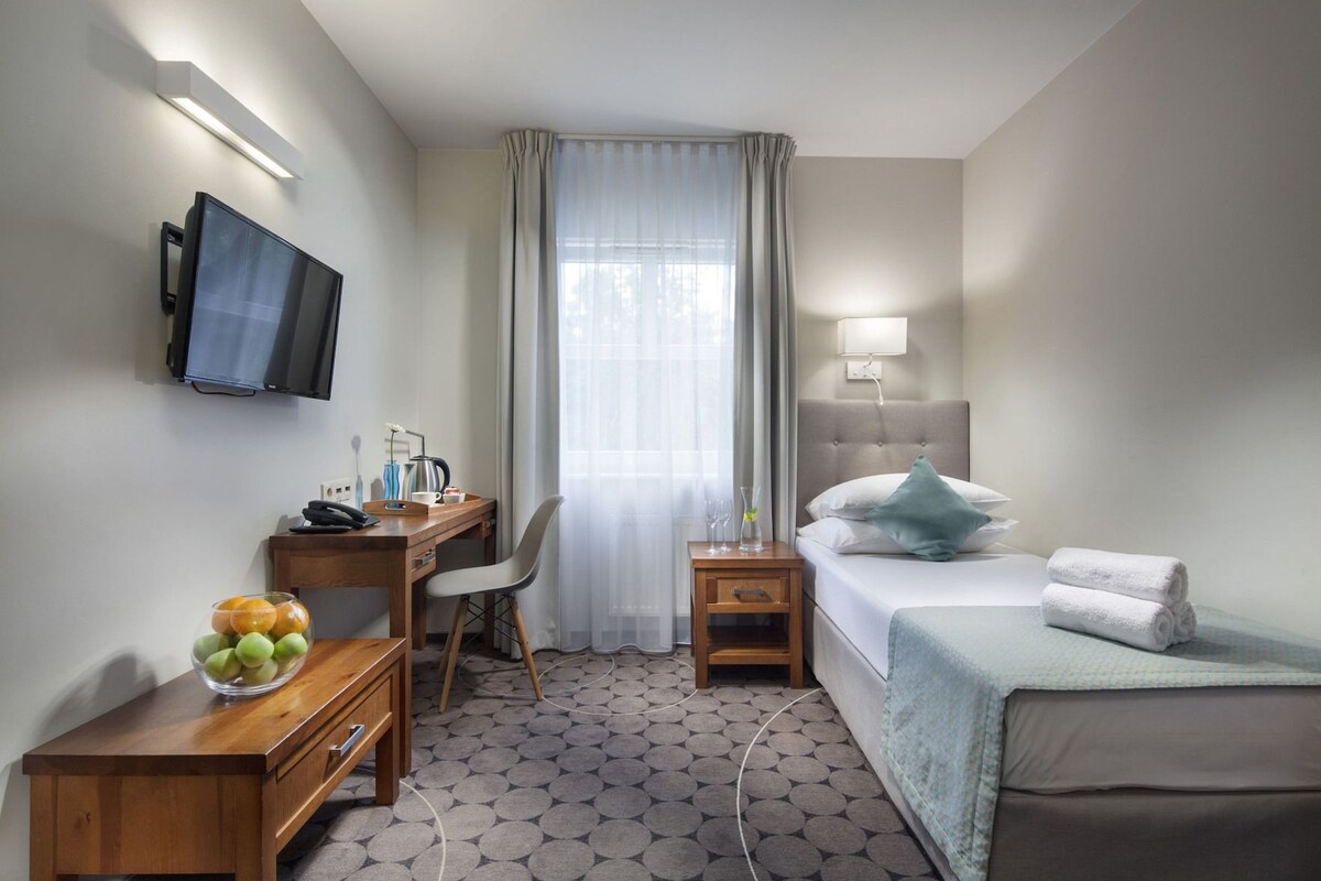 A hotel room with a bed, a desk, a chair, a TV, a bedside table with a lamp, and a window with curtains.