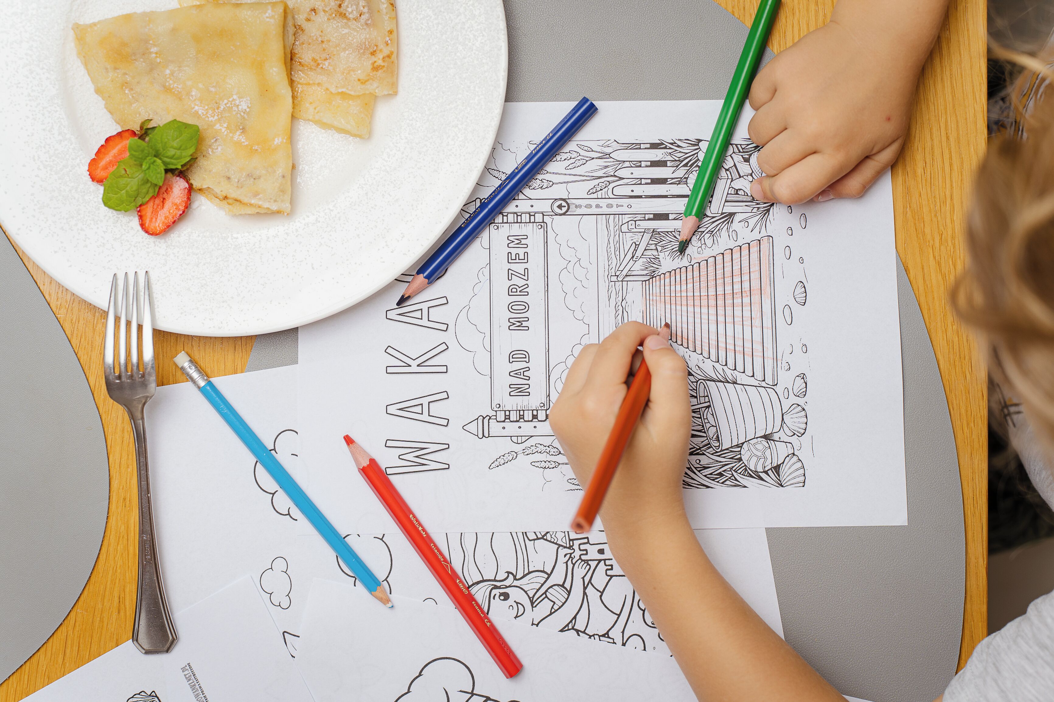 Un niño coloreando con un lápiz, un plato con tortitas y fresas, y un tenedor sobre una mesa.