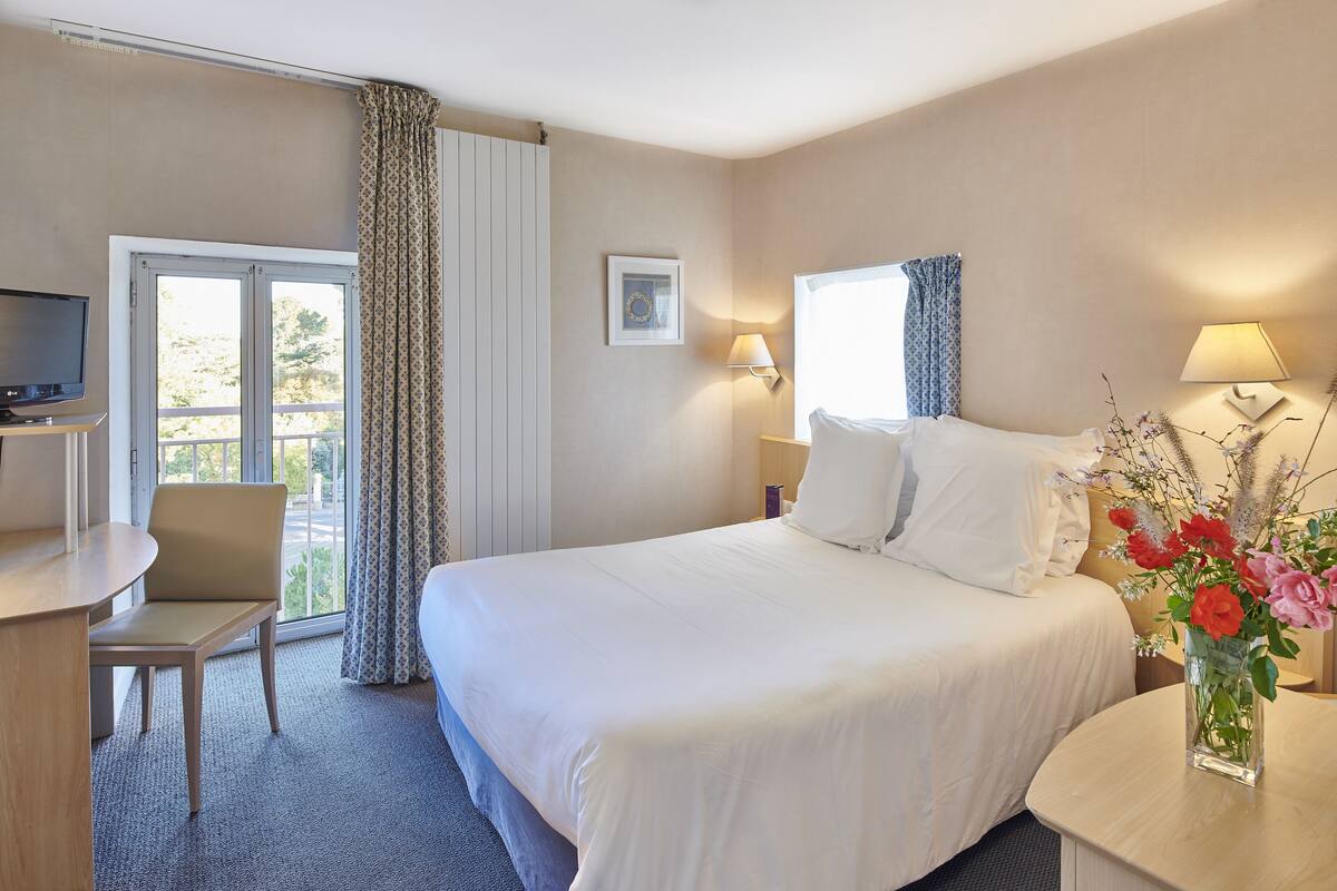 Une chambre d’hôtel comprenant un lit, un bureau, une chaise, une télévision, une fenêtre avec des rideaux et un vase avec des fleurs.