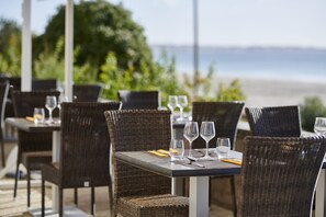 A dining area with wicker chairs and tables, each set with cutlery and wine glasses, overlooking a beach.