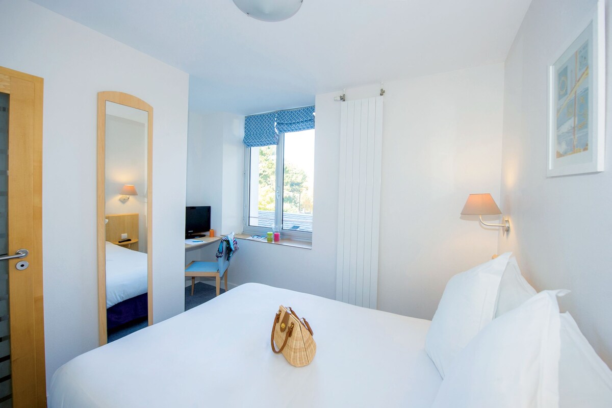 Habitación de hotel con cama, escritorio, silla, lámpara, ventana con cortinas y puerta.